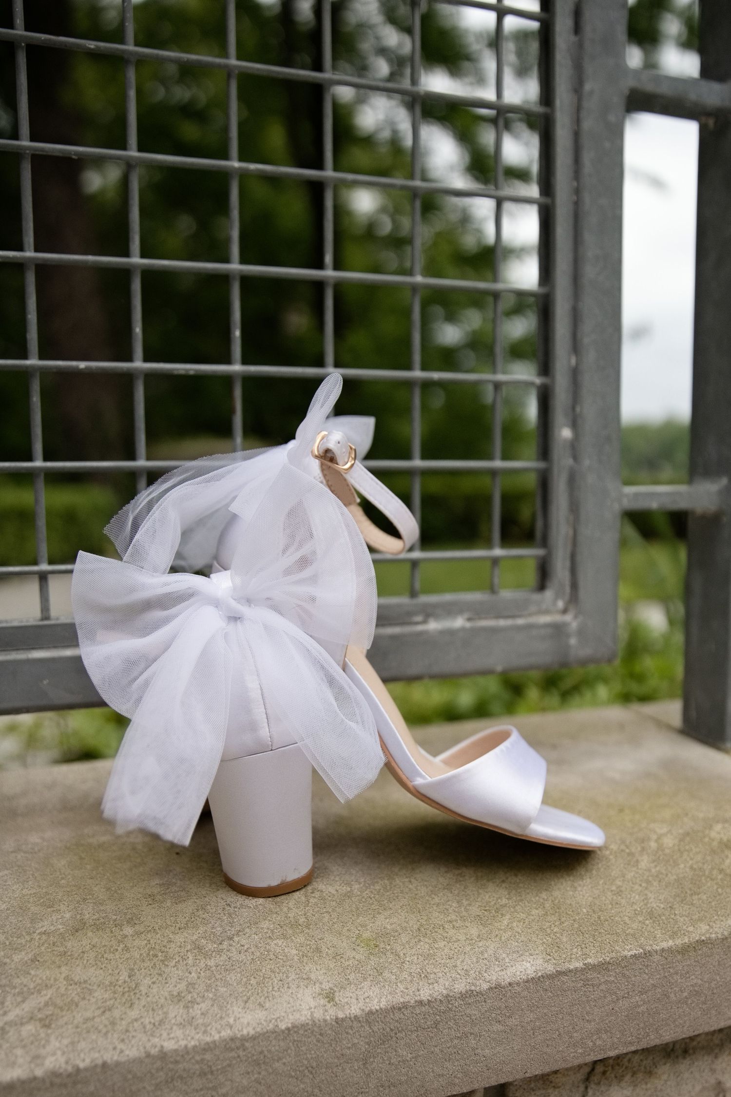 White bridal heels with tulle bow detail displayed on stone ledge near metal gate outdoors.