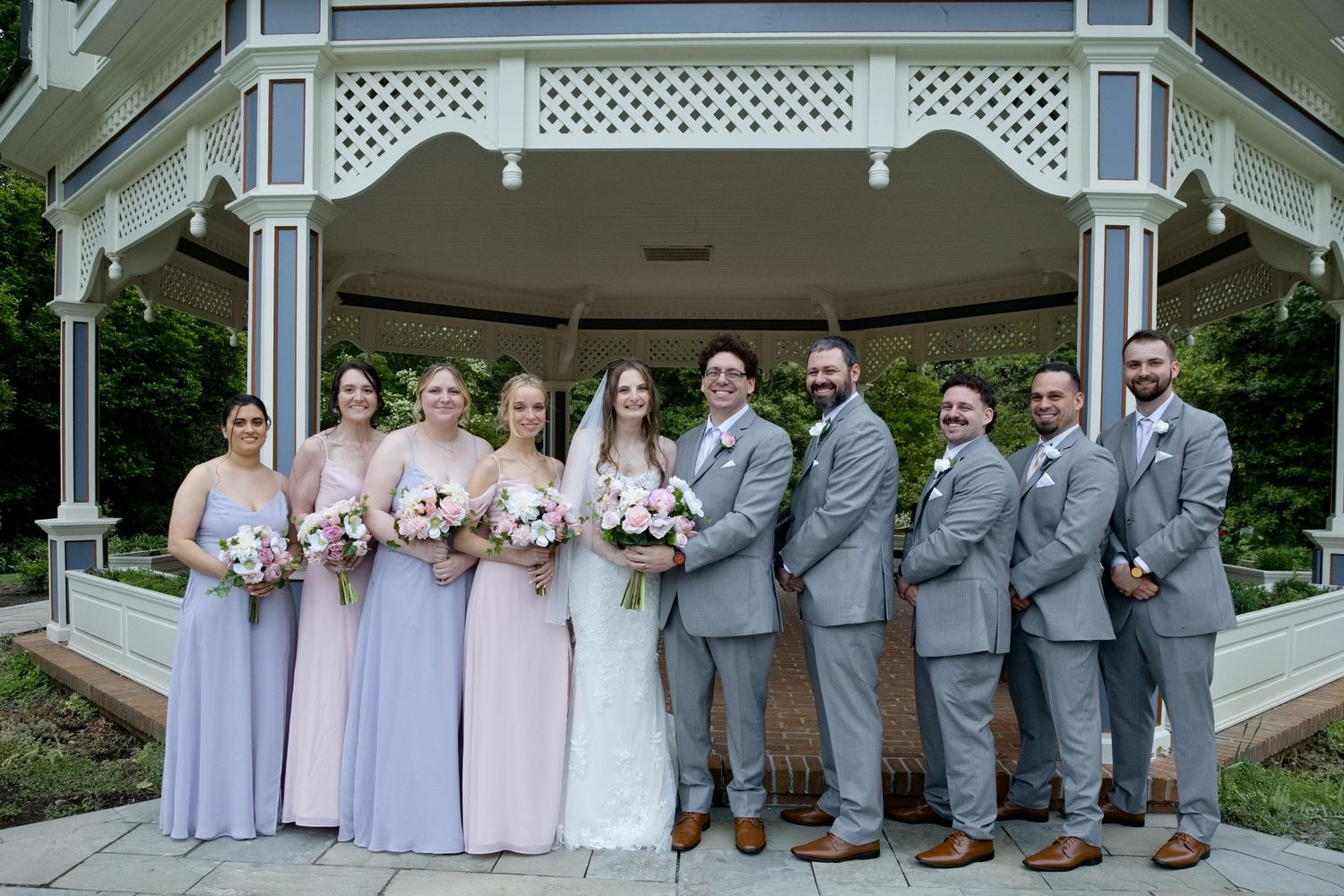 Wedding party poses before a gazebo, bridesmaids in lavender gowns and groomsmen in gray suits with bride and groom.