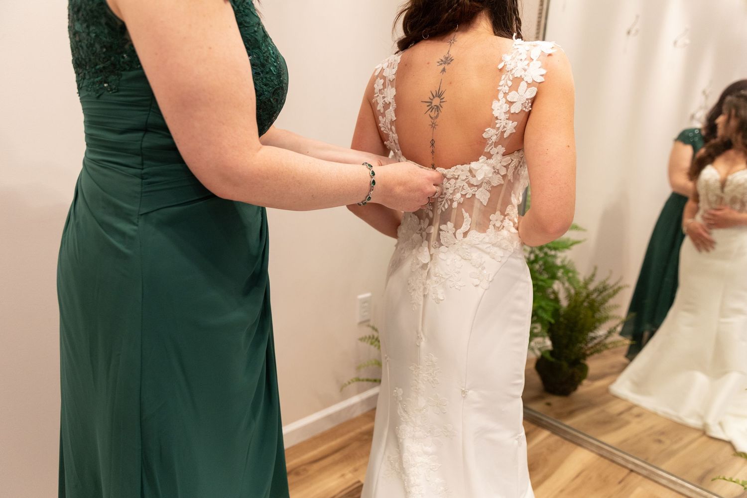 Woman in green dress buttoning the back of a lace wedding gown on a bride in a bridal boutique.