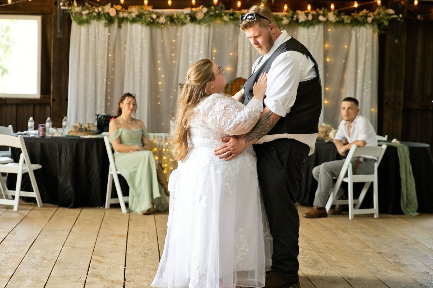 Bride and groom share their first dance in a rustic barn wedding venue with string lights and floral backdrop.