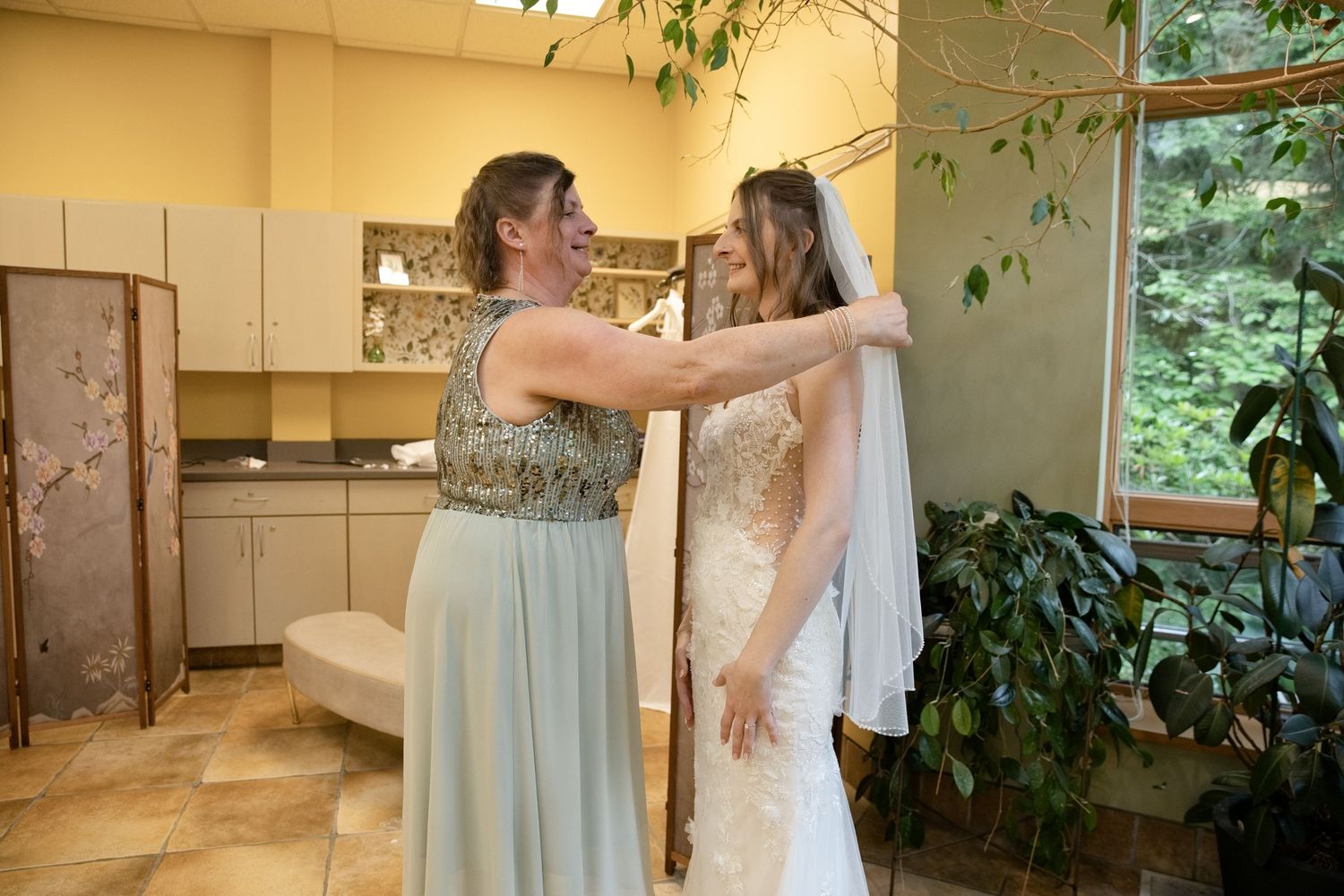 Bridesmaid in sage green dress adjusting bride's veil in elegant bridal suite before wedding ceremony.