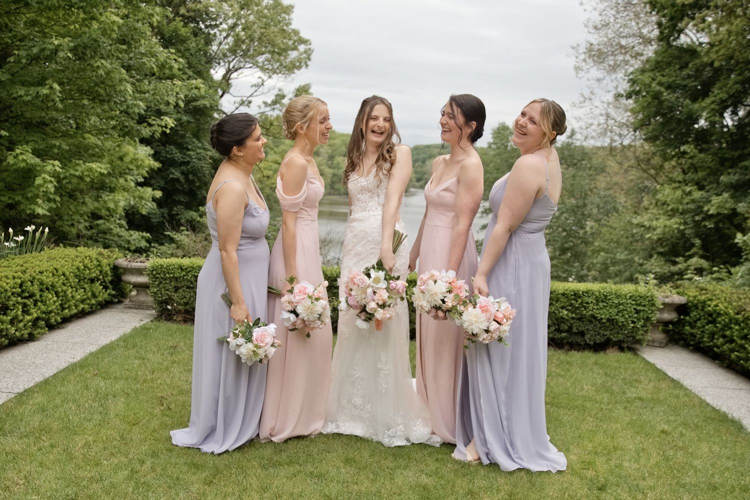Bride in white lace gown poses with four bridesmaids in lavender dresses holding pink bouquets in a garden.