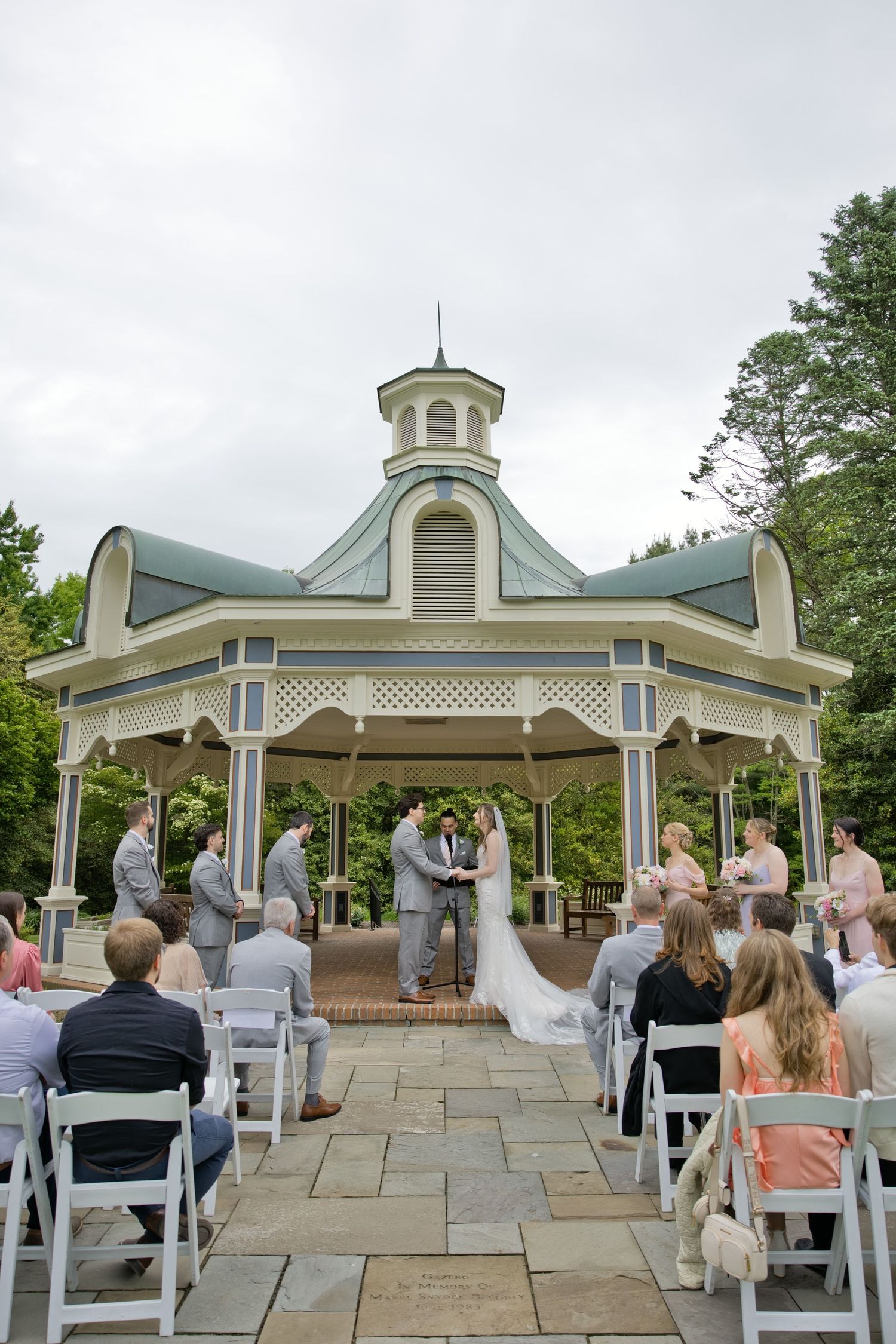 Couple exchanges vows under elegant gazebo surrounded by wedding party and seated guests outdoors.
