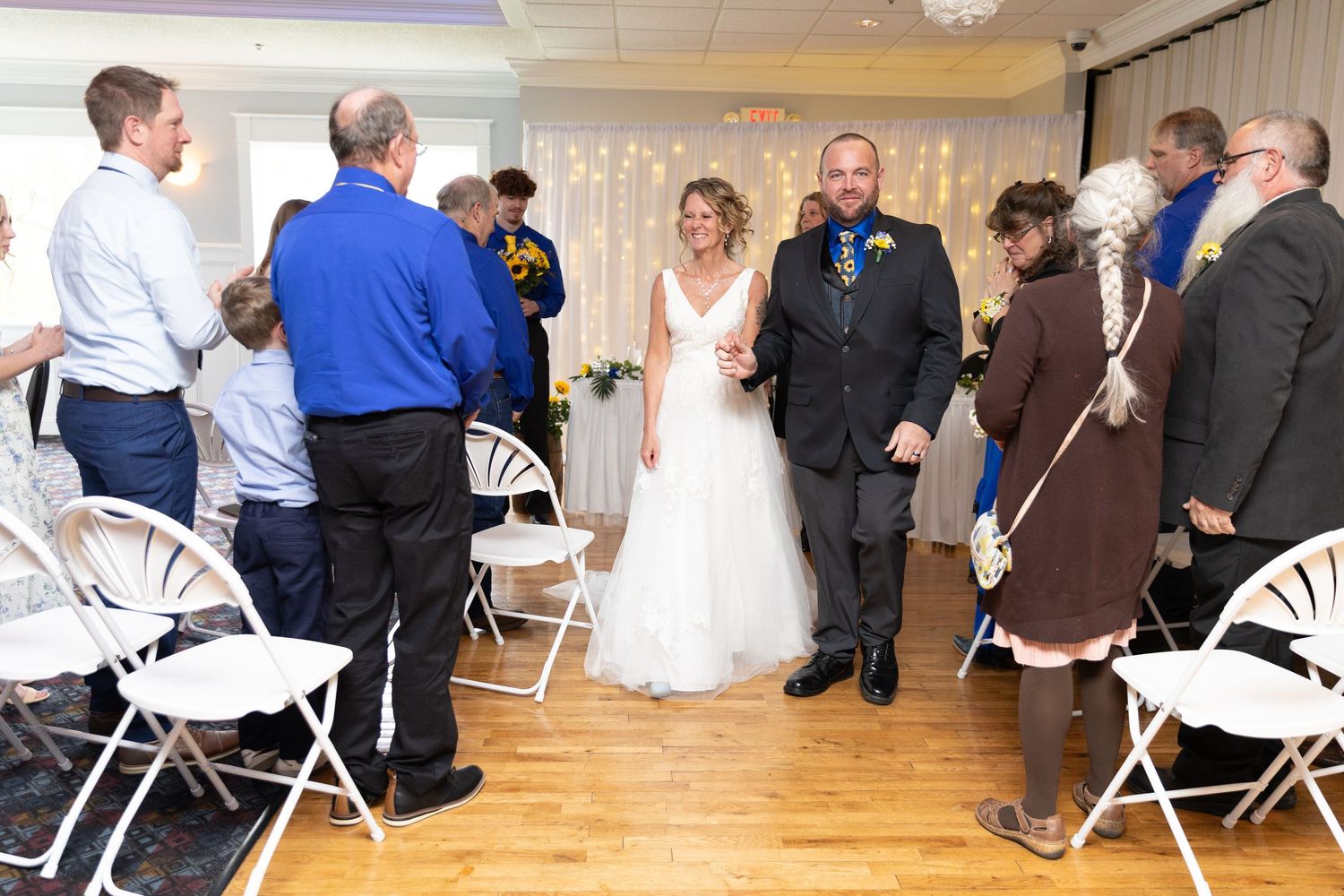 Bride and groom walk down the aisle at indoor wedding ceremony with guests applauding.