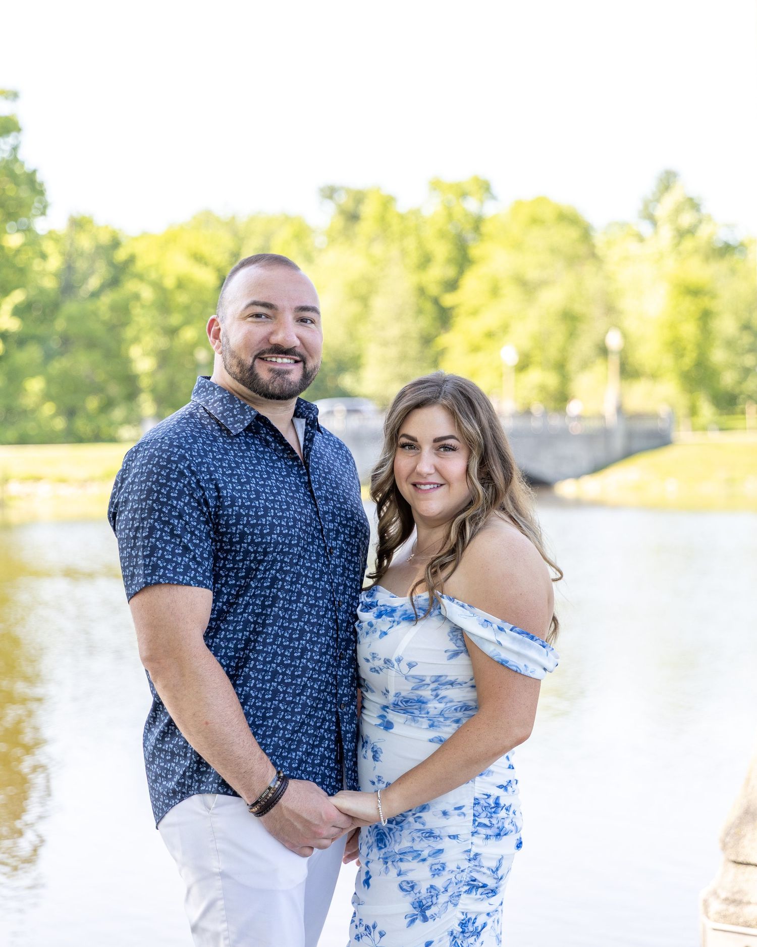 Smiling couple posing together near a serene lake with a bridge visible in the background.