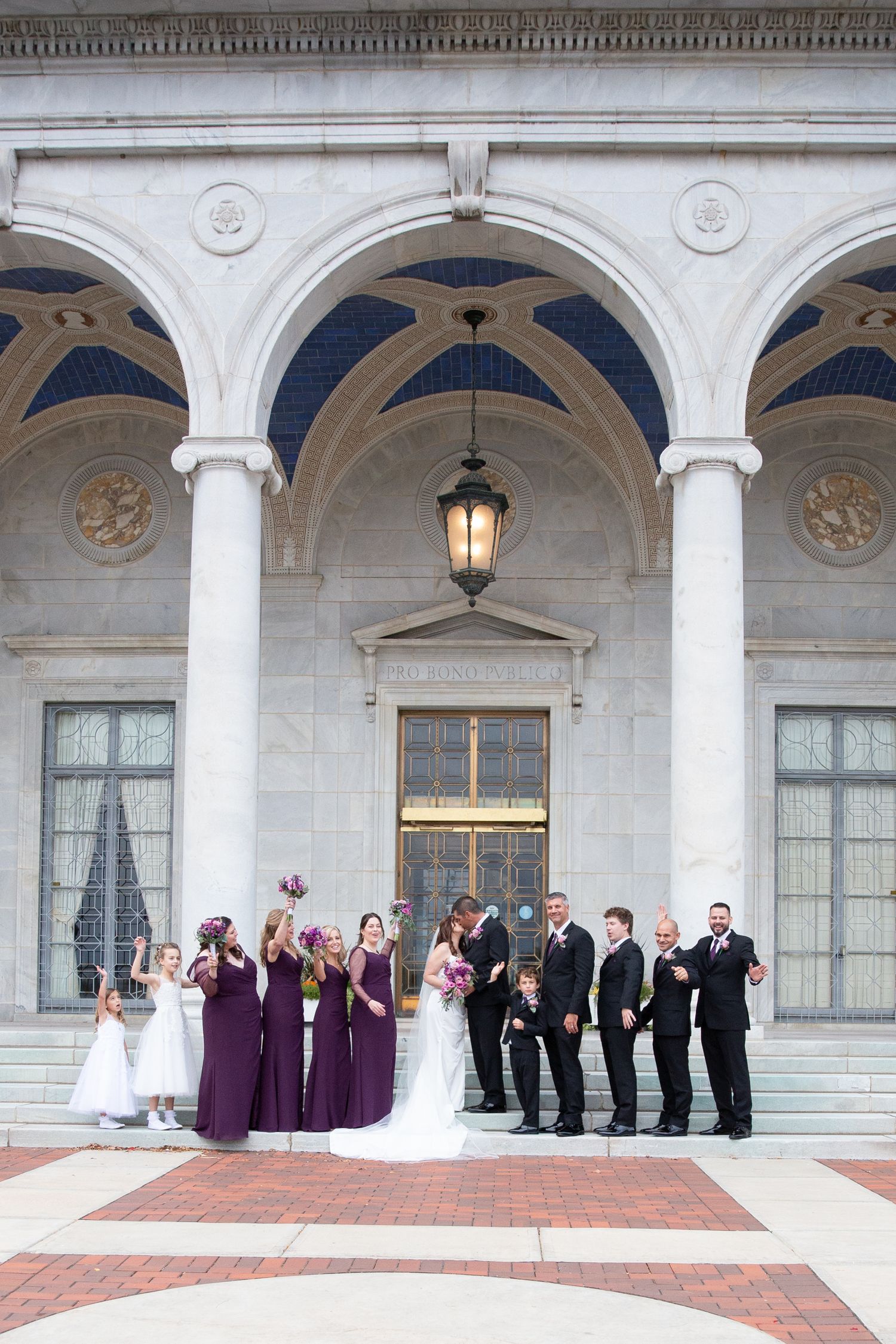 Wedding party lined up under classical white stone archway with columns.