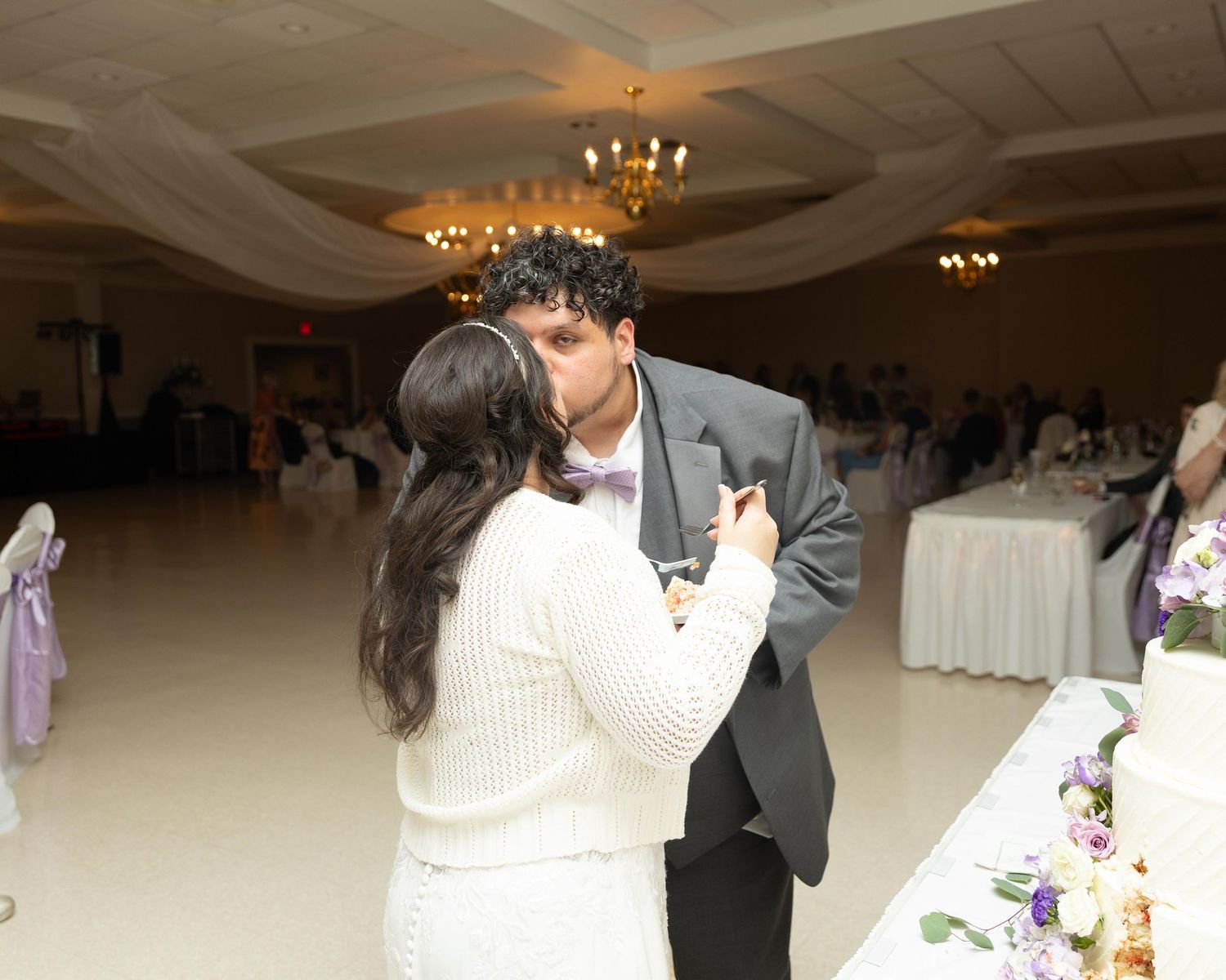Couple sharing a kiss at wedding reception near decorated cake table in elegant ballroom venue.