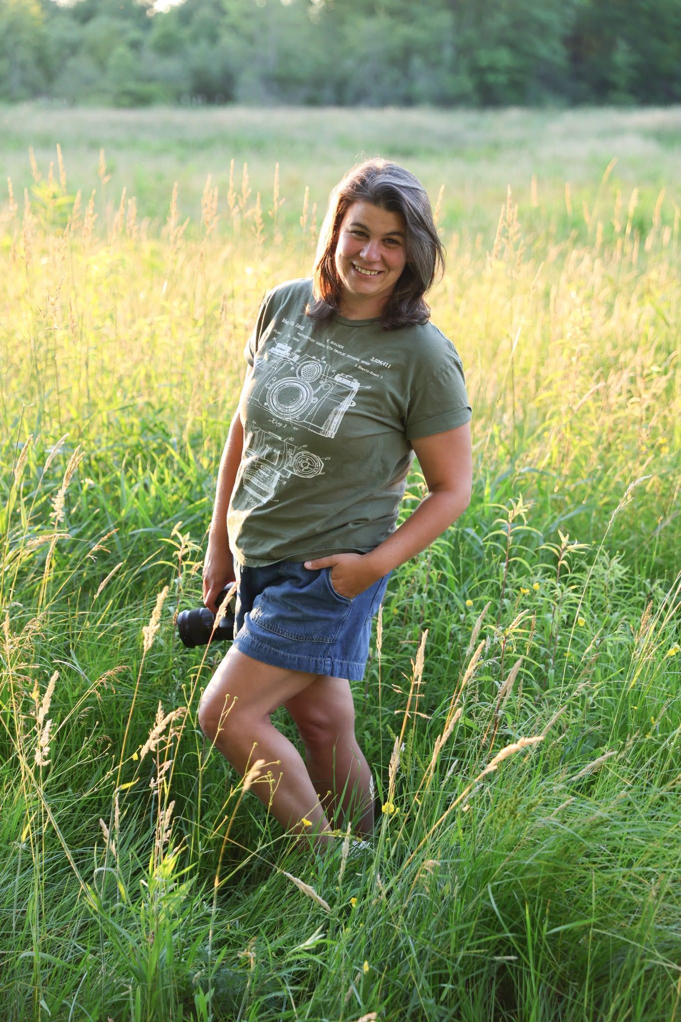 Smiling young woman in green t-shirt and denim shorts holding camera while standing in tall grass field at golden hour.