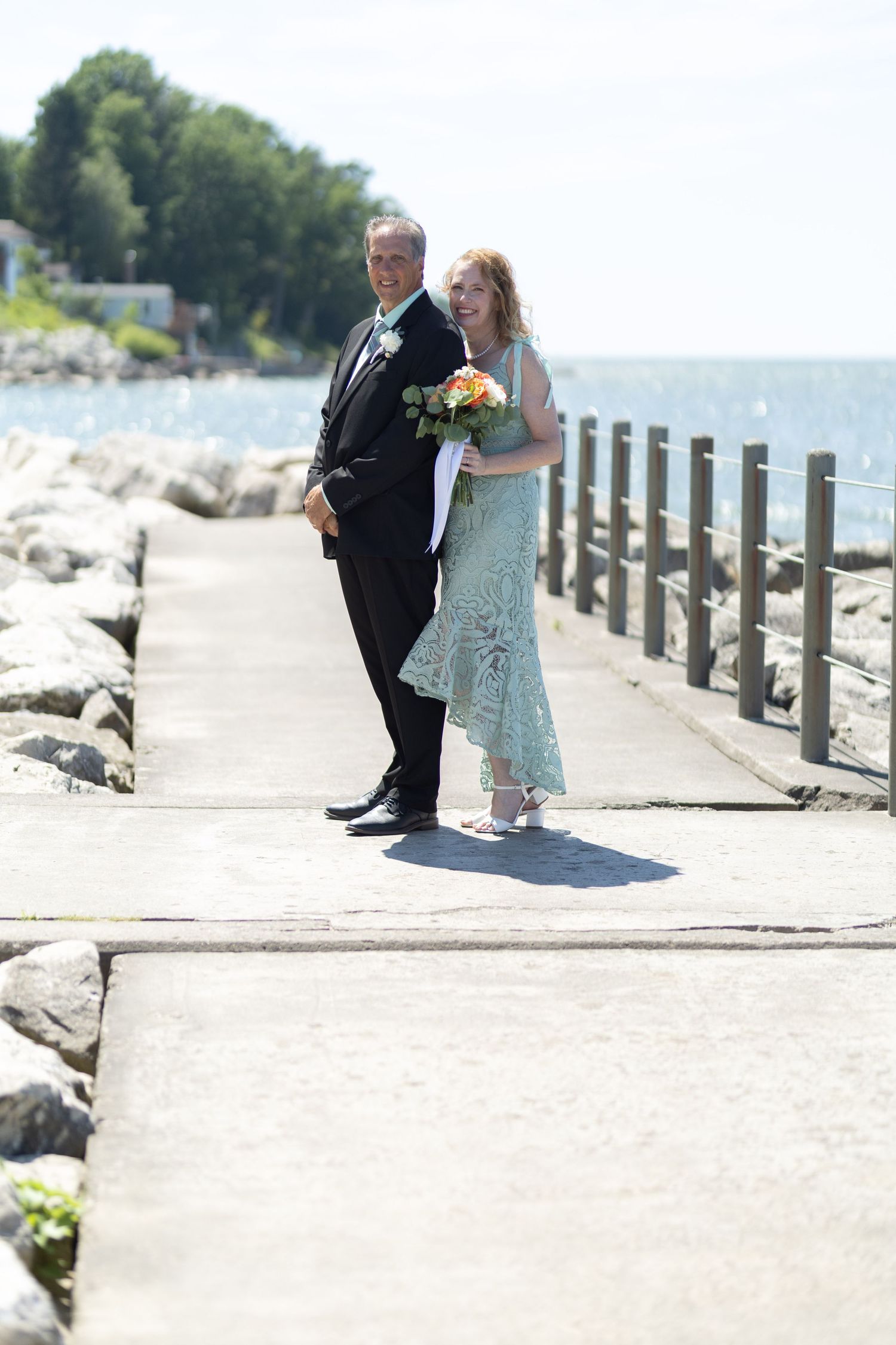 A couple in formal attire share an intimate moment on a beachfront walkway with wooden posts.