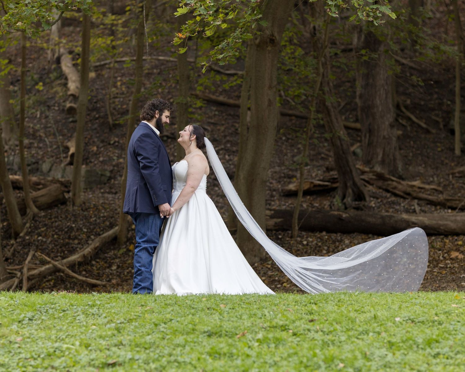Bride in flowing white gown with long veil poses with groom in woodland setting.