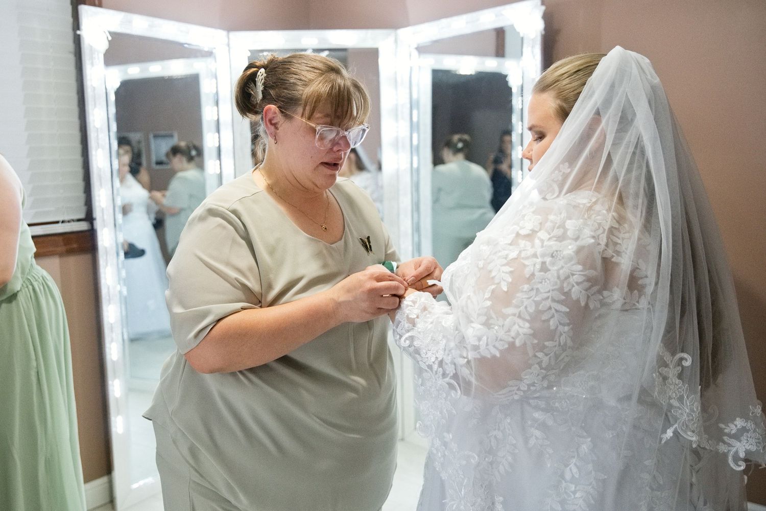 A bridal consultant helps a bride in a lace wedding dress and veil during a fitting in a dressing room.