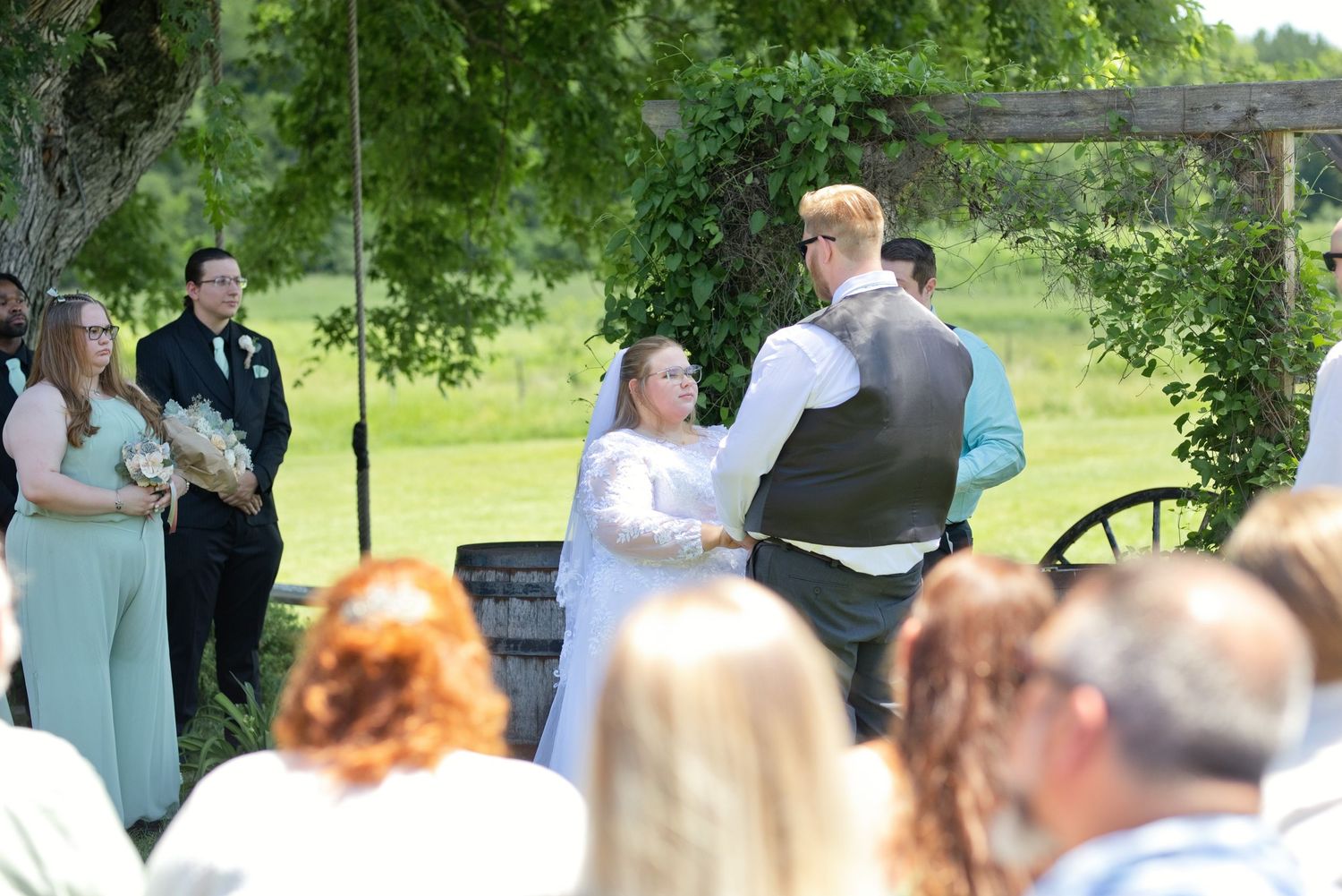 Outdoor wedding ceremony with bride and groom holding hands at rustic wooden arch, surrounded by guests in green field.