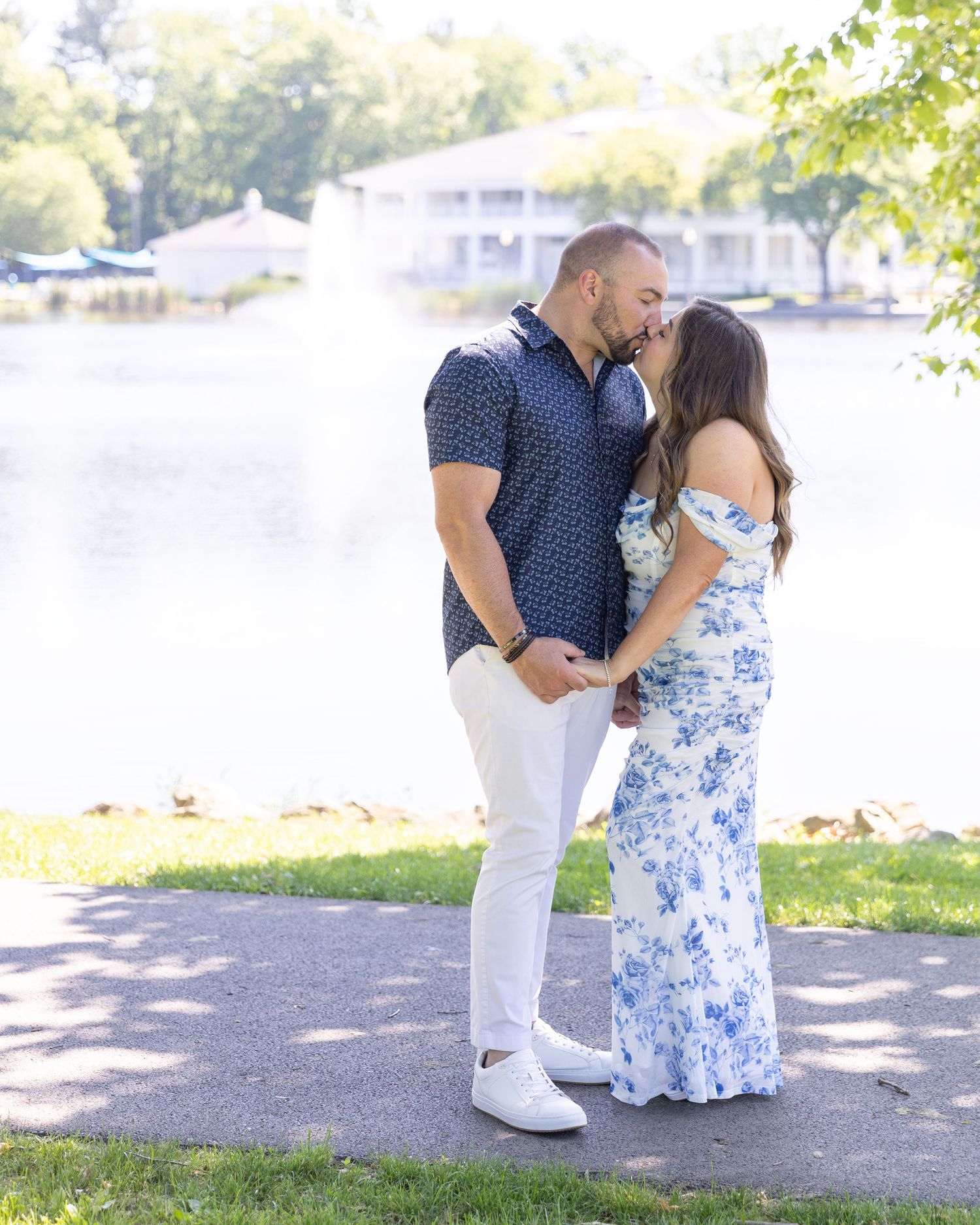 Couple sharing a romantic kiss by a serene lake, man in blue shirt and woman in floral dress, lush greenery background.