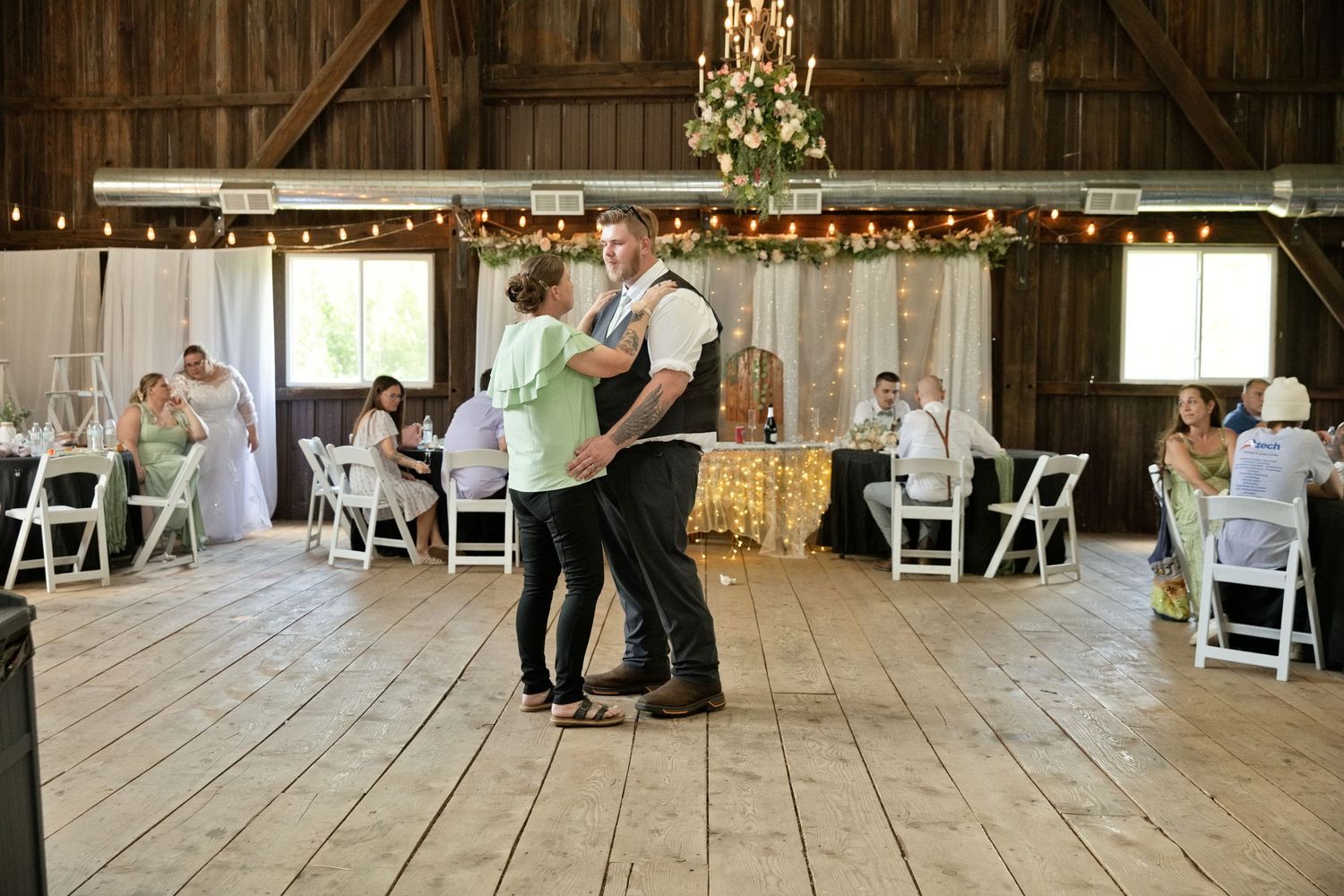 Couple sharing first dance in rustic barn wedding venue with string lights and white chairs.