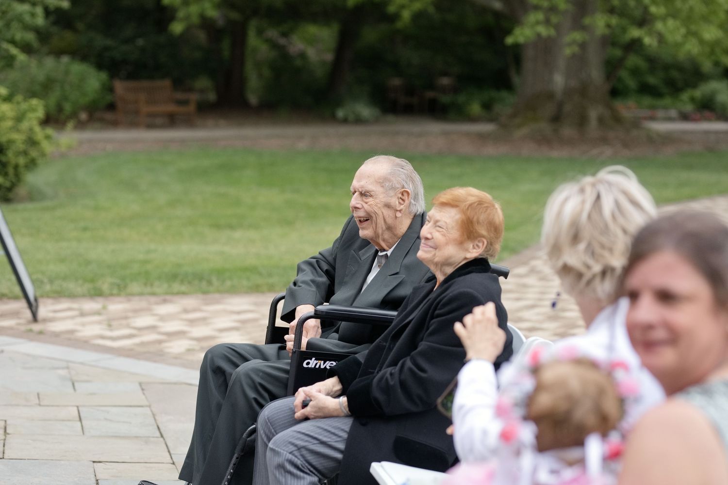 Elderly couple sitting together outdoors at a garden event, smiling warmly on a paved patio surrounded by greenery.
