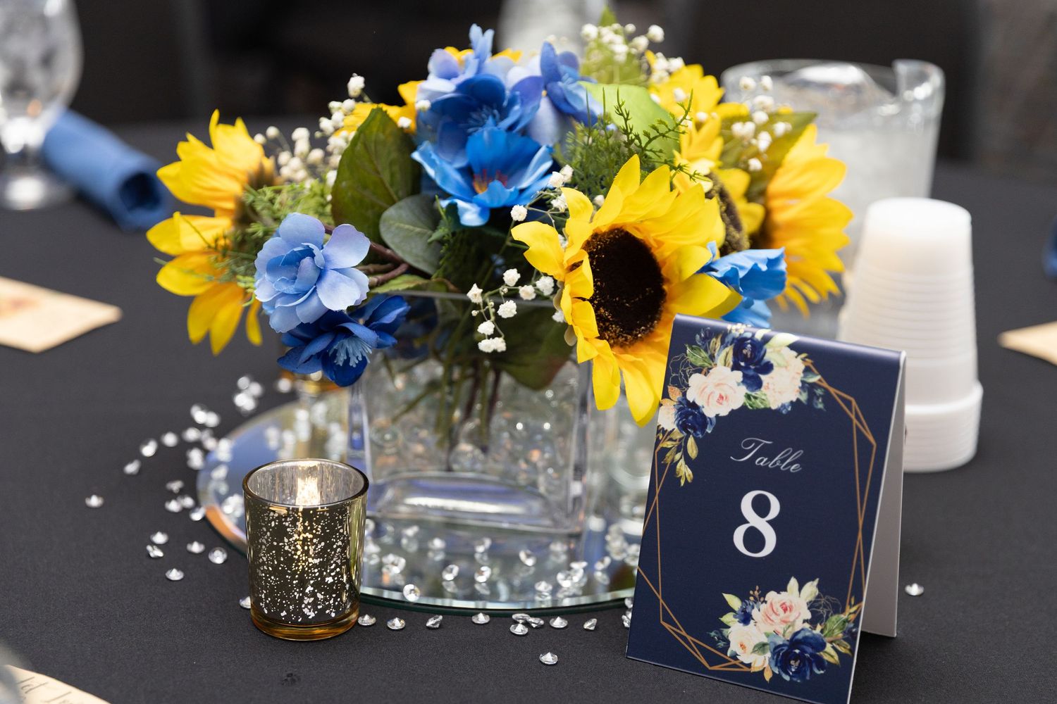 Blue and yellow sunflower wedding centerpiece with baby's breath in glass vase beside Table 8 sign on dark tablecloth.