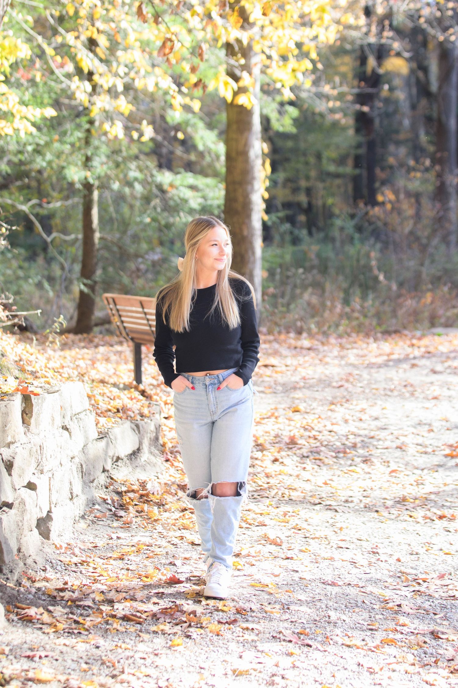 Young woman in a black shirt walking, smiling, with trees and sunlight in the background.