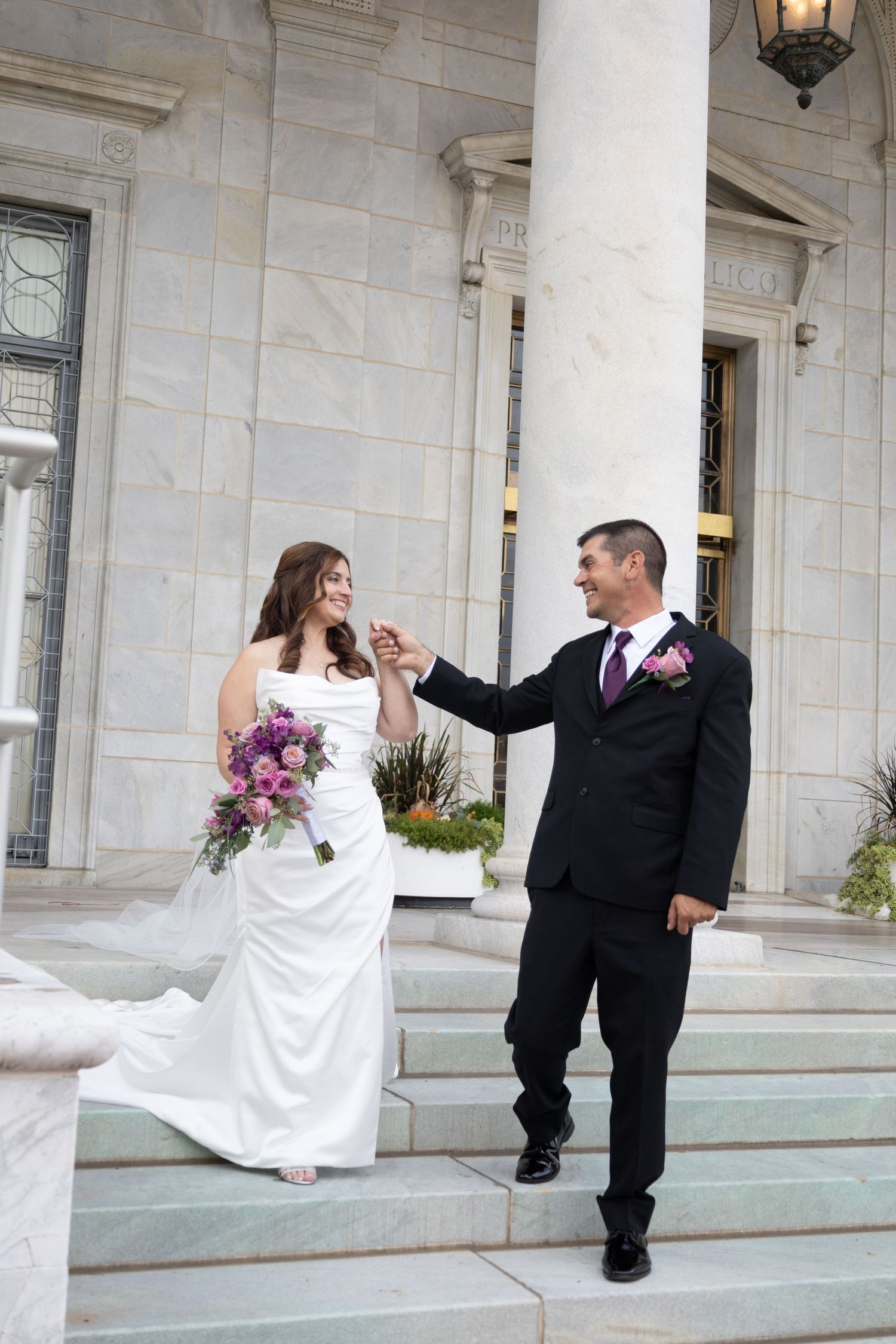 Playful wedding couple poses on white stone steps outside building.