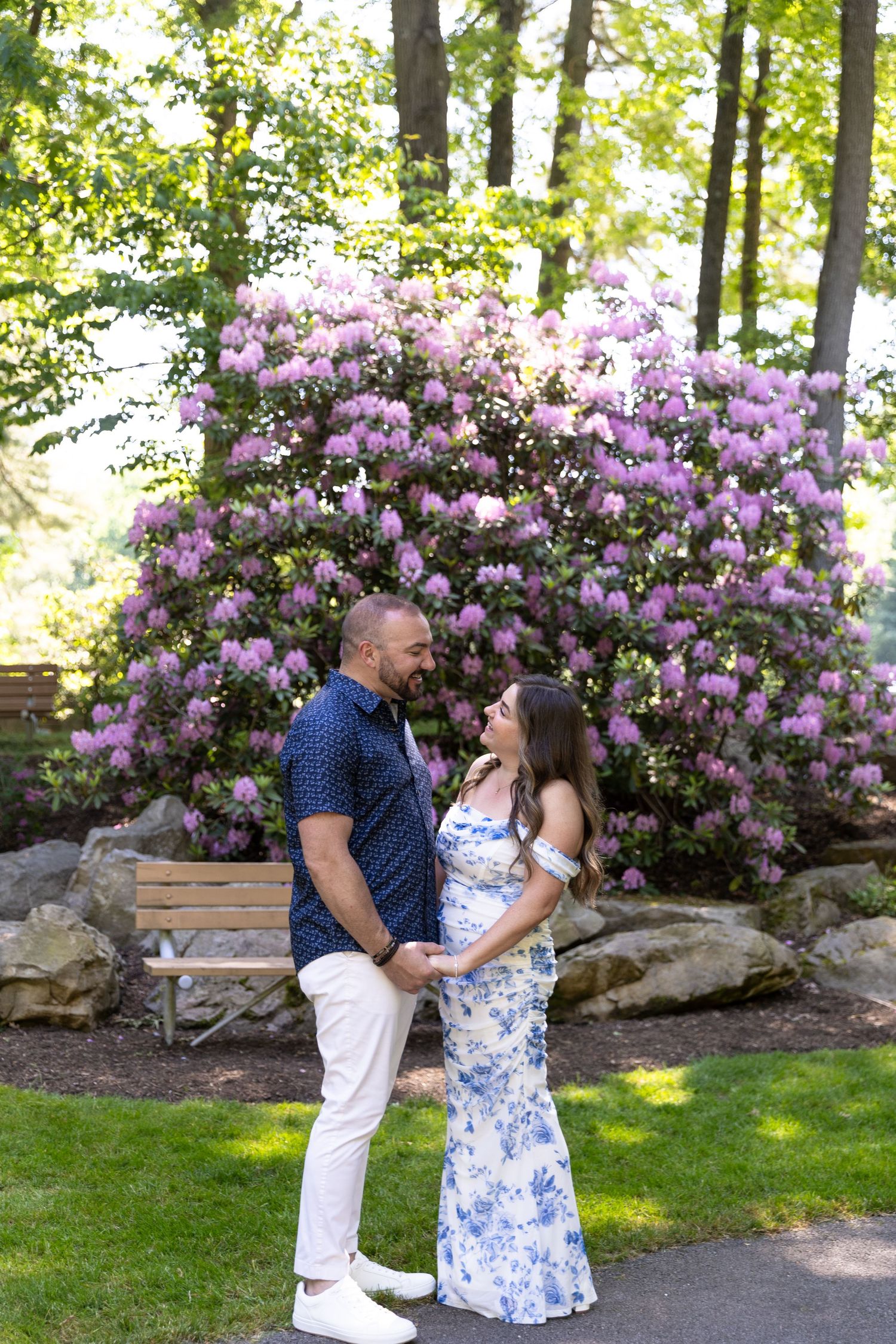 Couple holding hands and gazing at each other in front of blooming purple rhododendrons in a lush garden.
