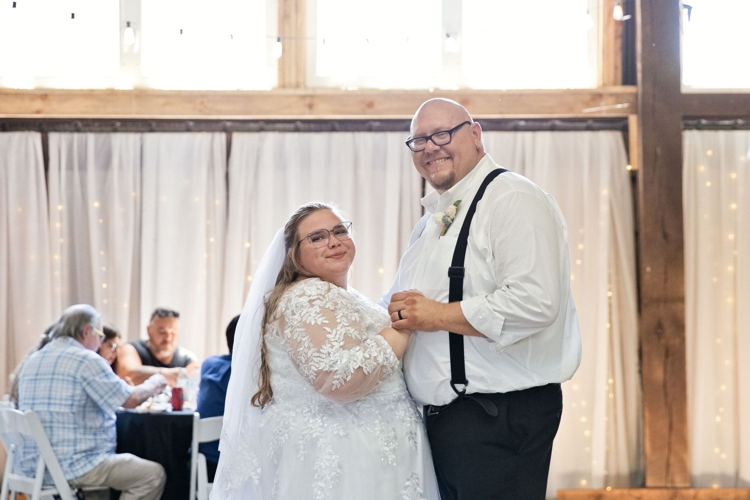 Bride and groom share first dance in rustic barn wedding venue, bride in lace gown with veil, groom in suspenders.