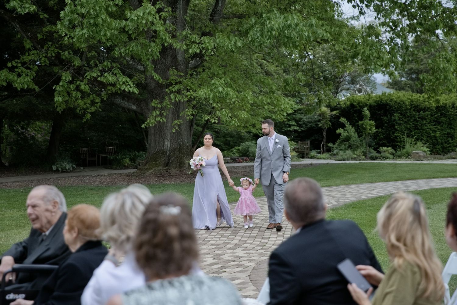 Wedding party walks down outdoor aisle, bridesmaid in lavender gown with flower girl in pink dress and groomsman in gray suit.