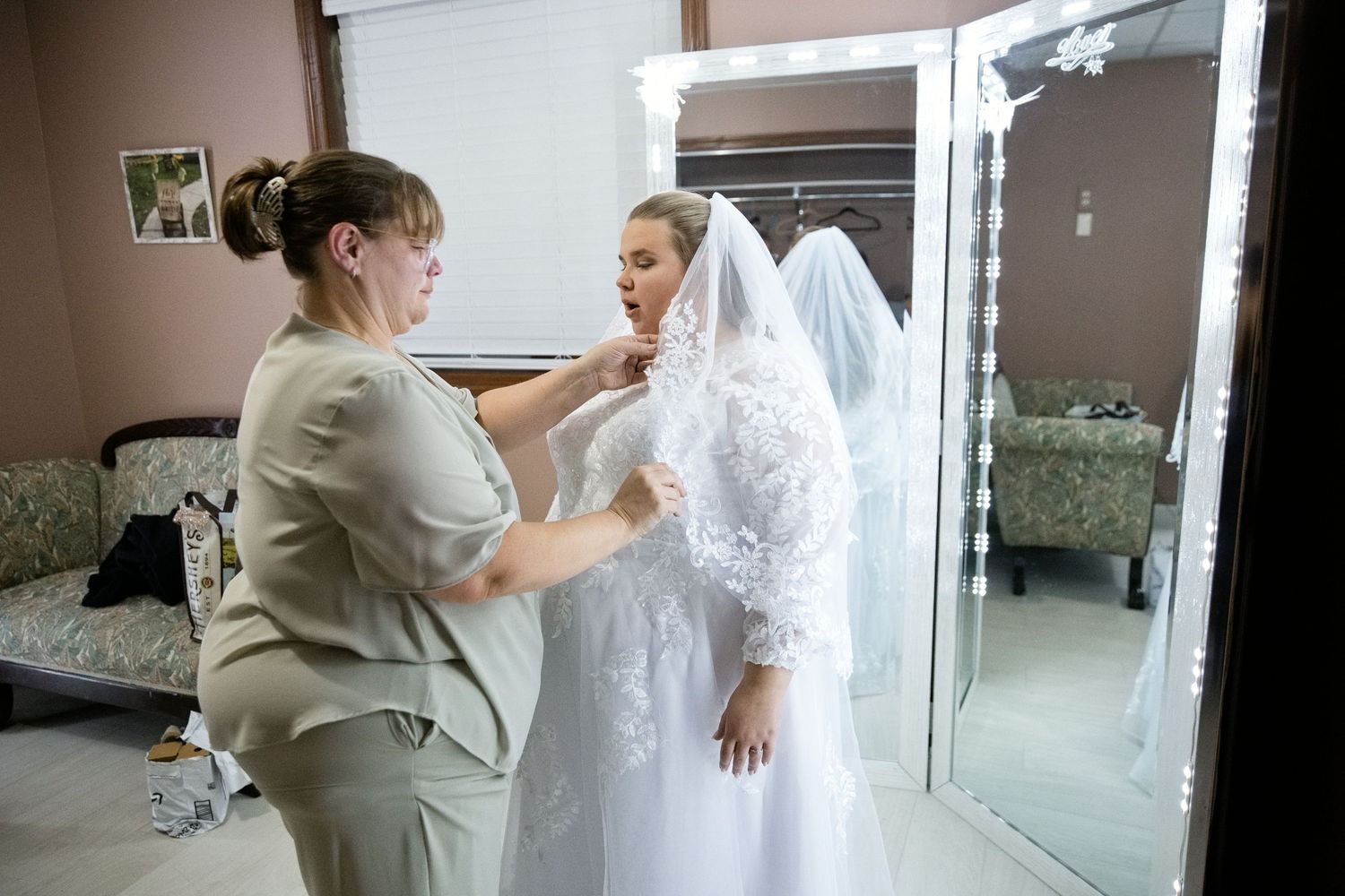A bride in a white wedding dress and veil being helped by a woman in a beige dress in a bridal suite.