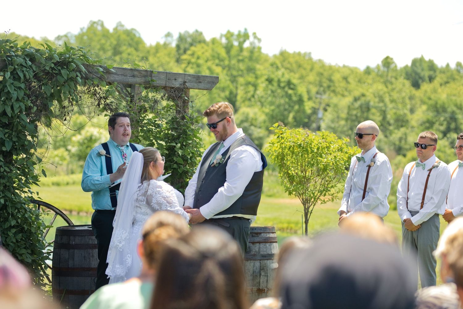 Couple exchanges vows at outdoor wedding ceremony with groomsmen and officiant near rustic wine barrels.