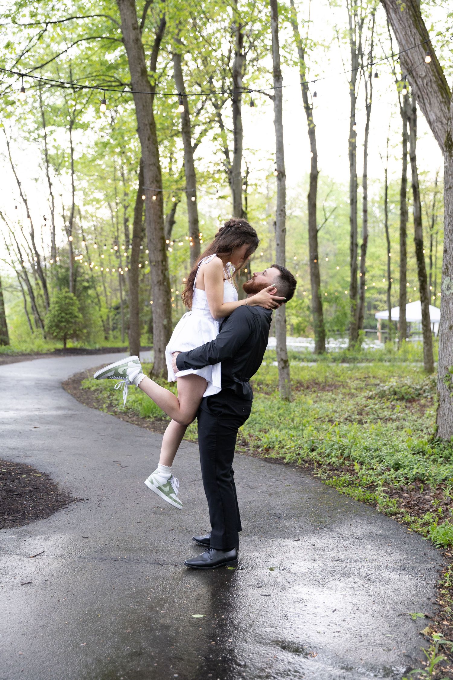 Couple sharing a romantic kiss in a lush green park, man lifting woman in white dress on a rainy day.