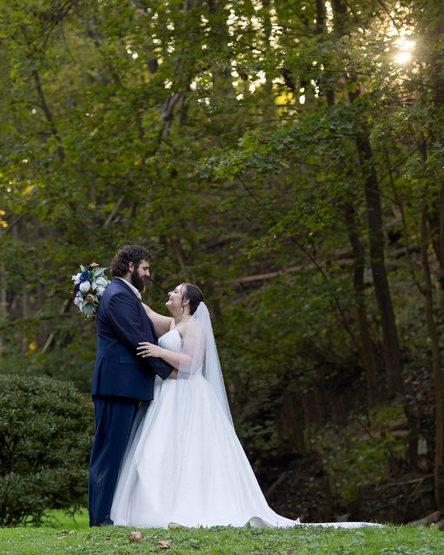 Romantic wedding portrait of couple embracing in wooded setting with sunlight filtering through trees.
