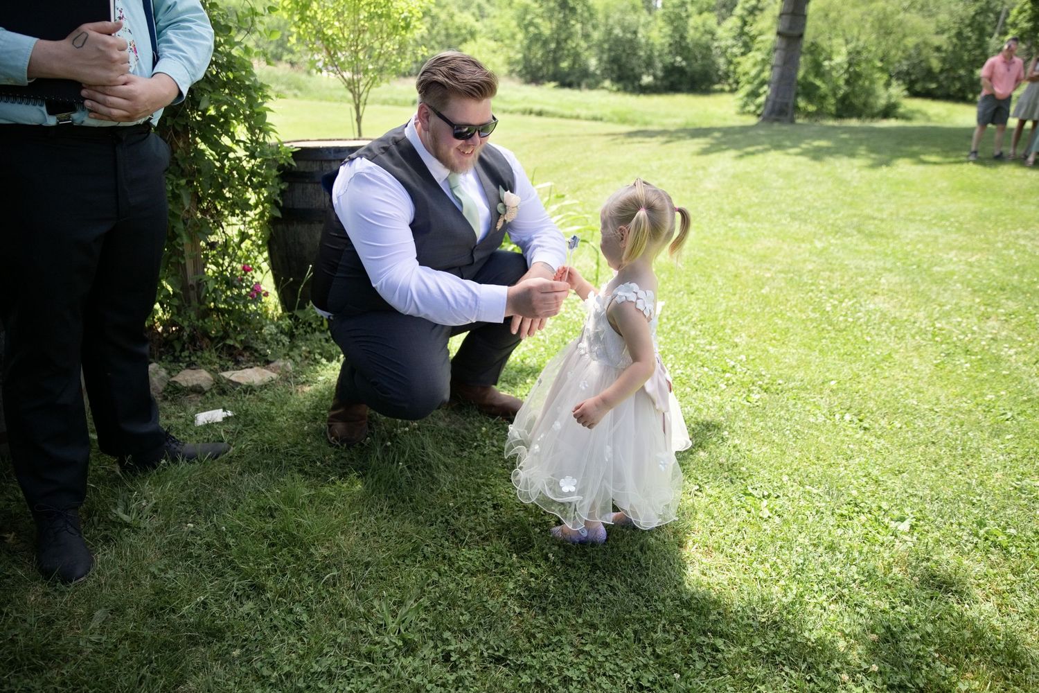 A groom in a vest and sunglasses kneels to interact with a young flower girl in a white dress on green grass.