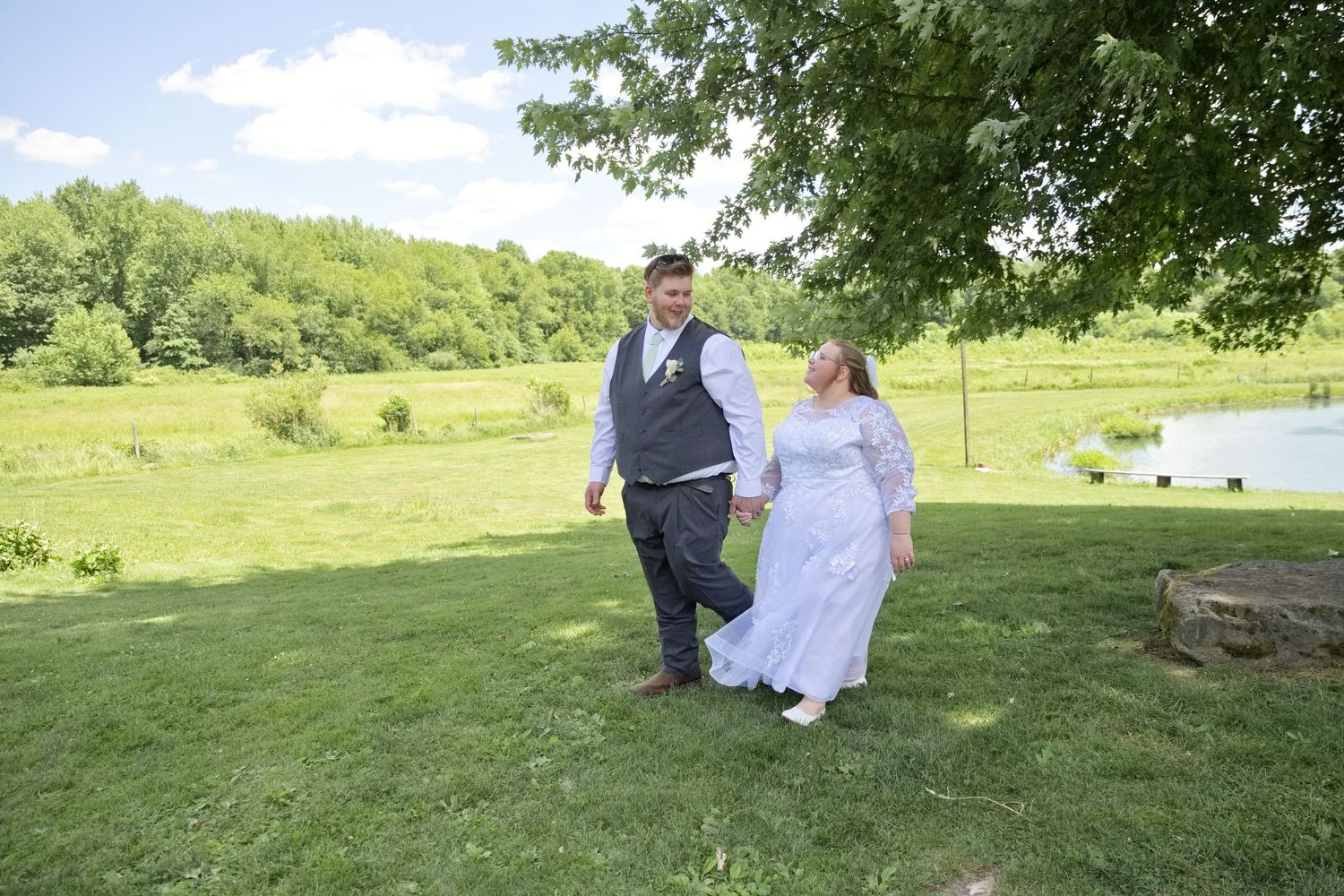 Bride and groom walking hand in hand on lush green lawn with pond and trees in background on wedding day.