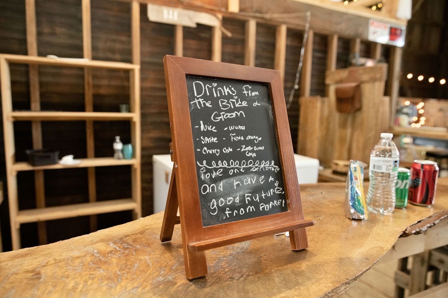 Chalkboard sign listing drinks for the bride and groom displayed on a wooden bar at a rustic wedding venue.