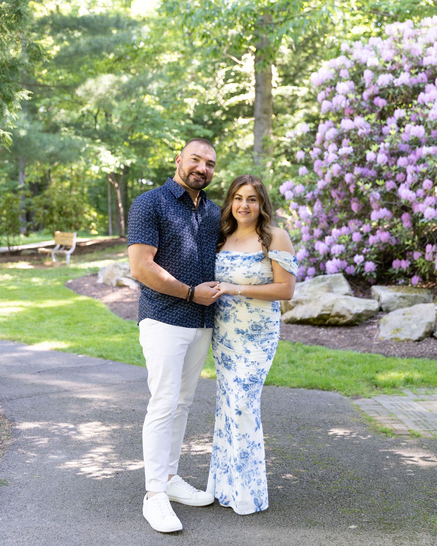 Couple posing together in a garden with blooming purple rhododendrons, woman in floral dress, man in navy shirt.