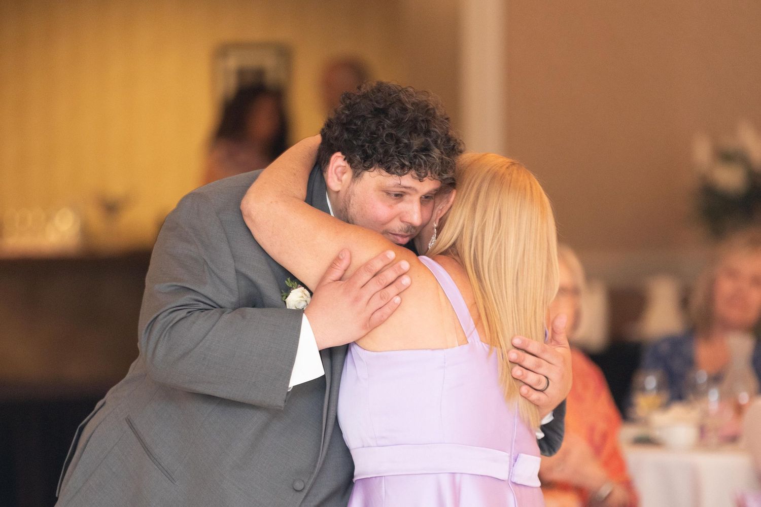 Groom in gray suit embraces bridesmaid in lavender dress at a wedding reception.