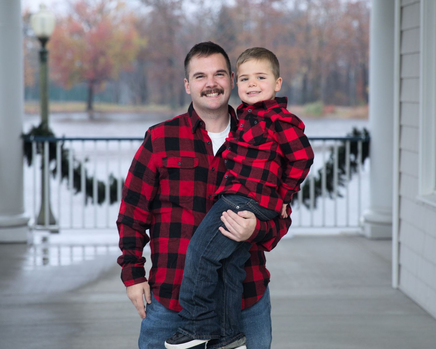 Family wearing coordinated red buffalo plaid flannel shirts pose on a white columned porch during autumn.