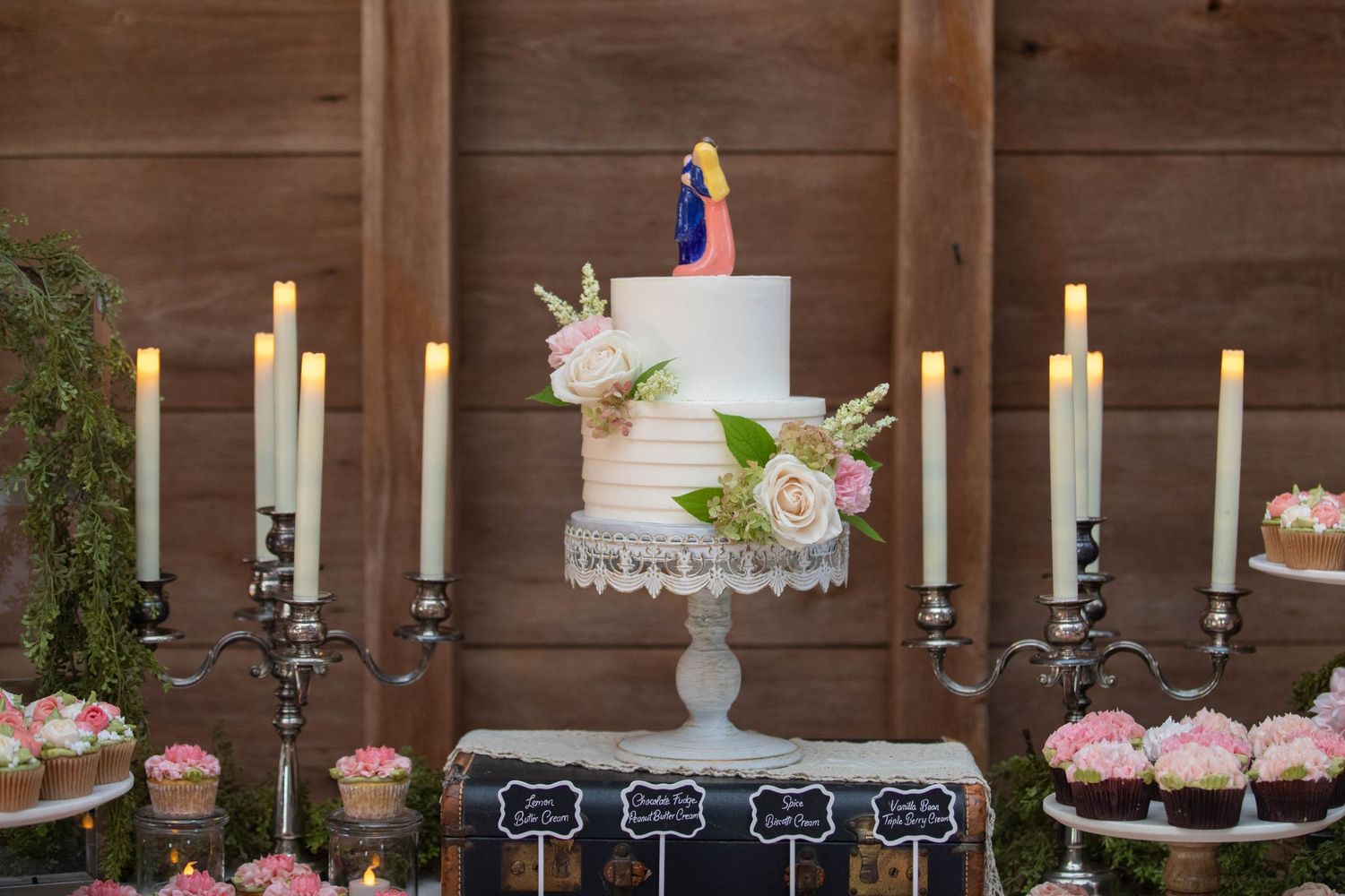 Wedding dessert table with white tiered cake, candelabras and pink cupcakes against wooden backdrop.