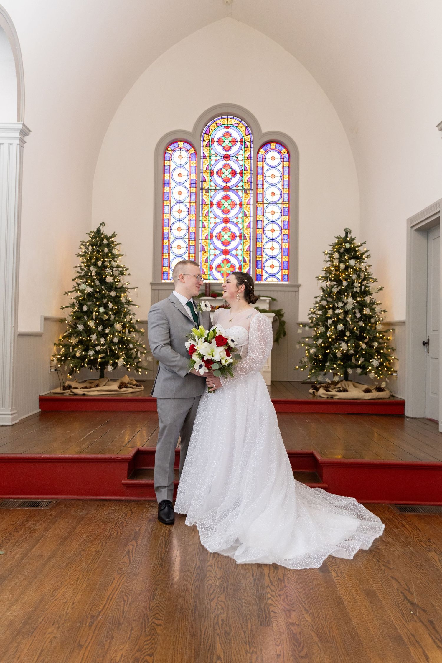 Bride and groom share a romantic moment in a church with stained glass windows and Christmas trees.