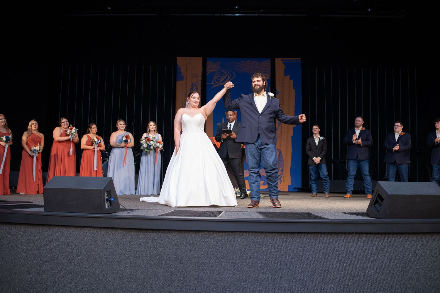 Newlyweds celebrate on stage during wedding ceremony with bridal party looking on.