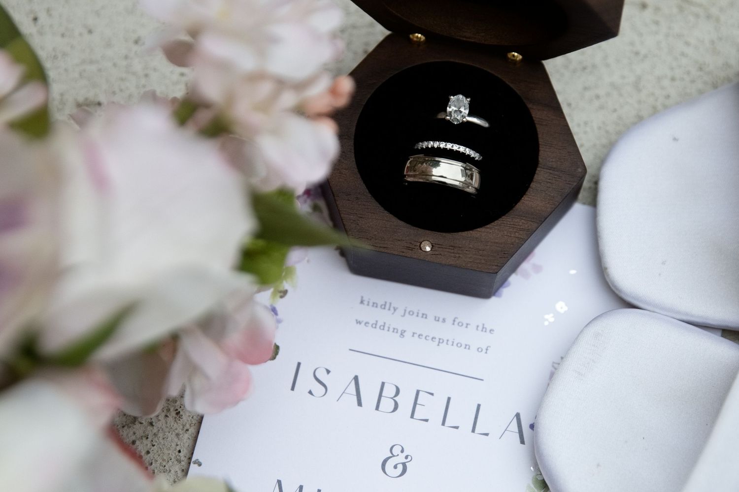 Wedding rings in a wooden box on an invitation for Isabella, with pink flowers nearby.