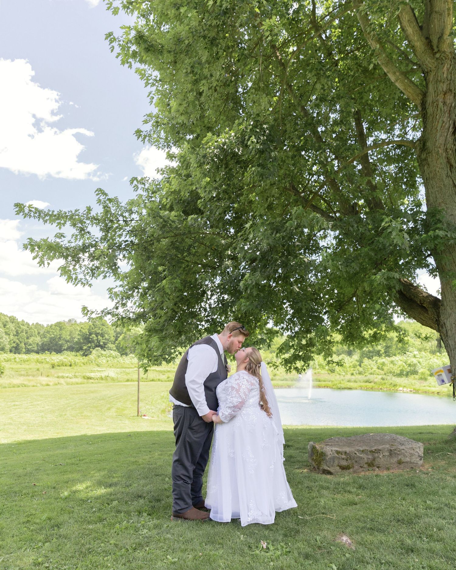 Bride and groom share a kiss under a large tree on a sunny day at an outdoor wedding venue with a pond.