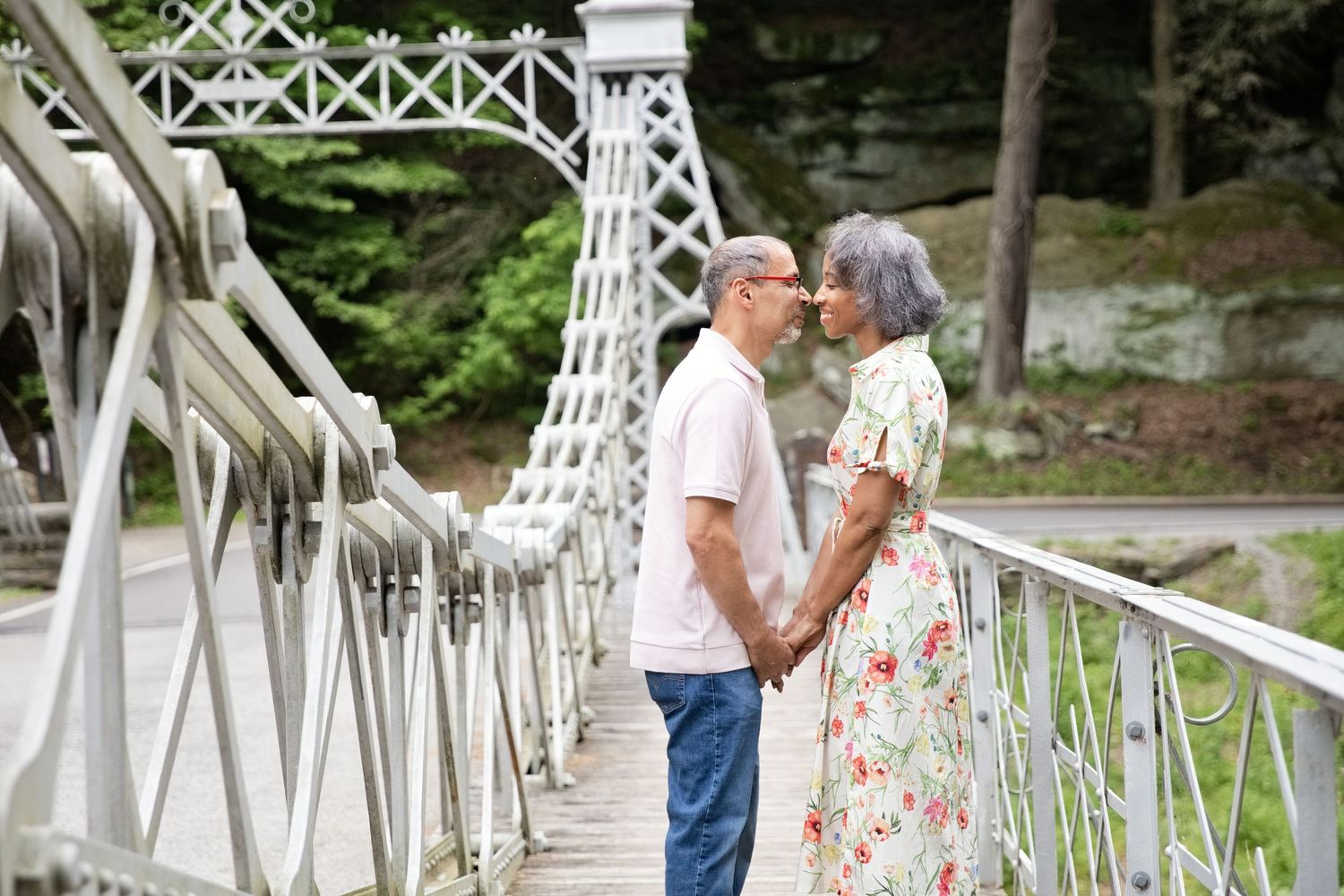 Elderly couple holding hands and facing each other on a white iron bridge surrounded by lush green trees.
