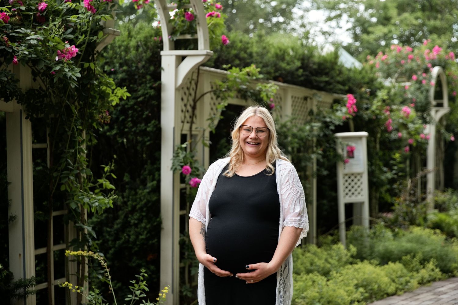 Pregnant woman in black dress and white cardigan smiling in a lush garden with pink roses and white arbors.