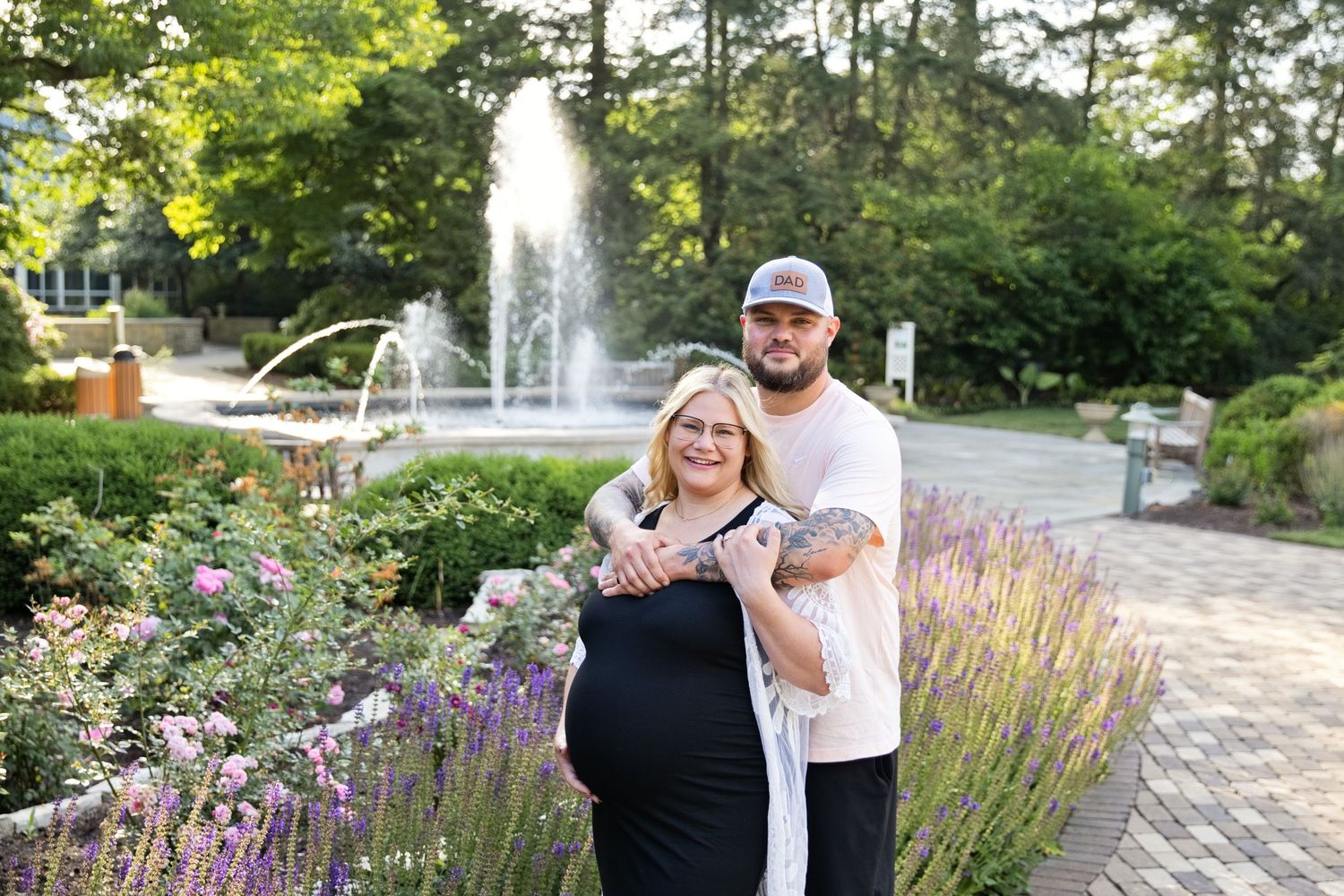 Pregnant couple posing in a garden with a fountain, woman in black dress, man in white shirt with tattoos.