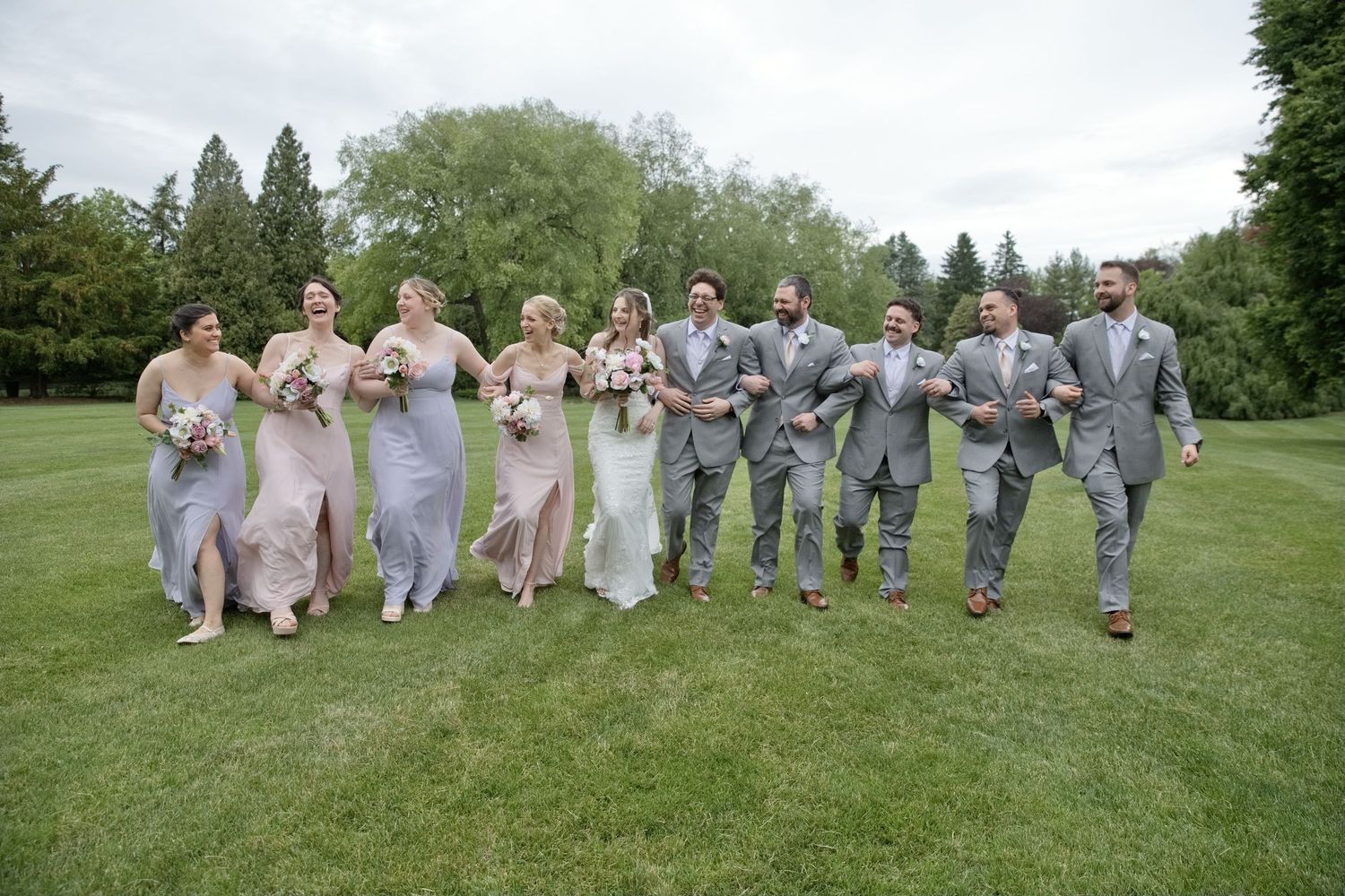 Wedding party walks on green lawn, bridesmaids in pink and gray gowns, groomsmen in gray suits.
