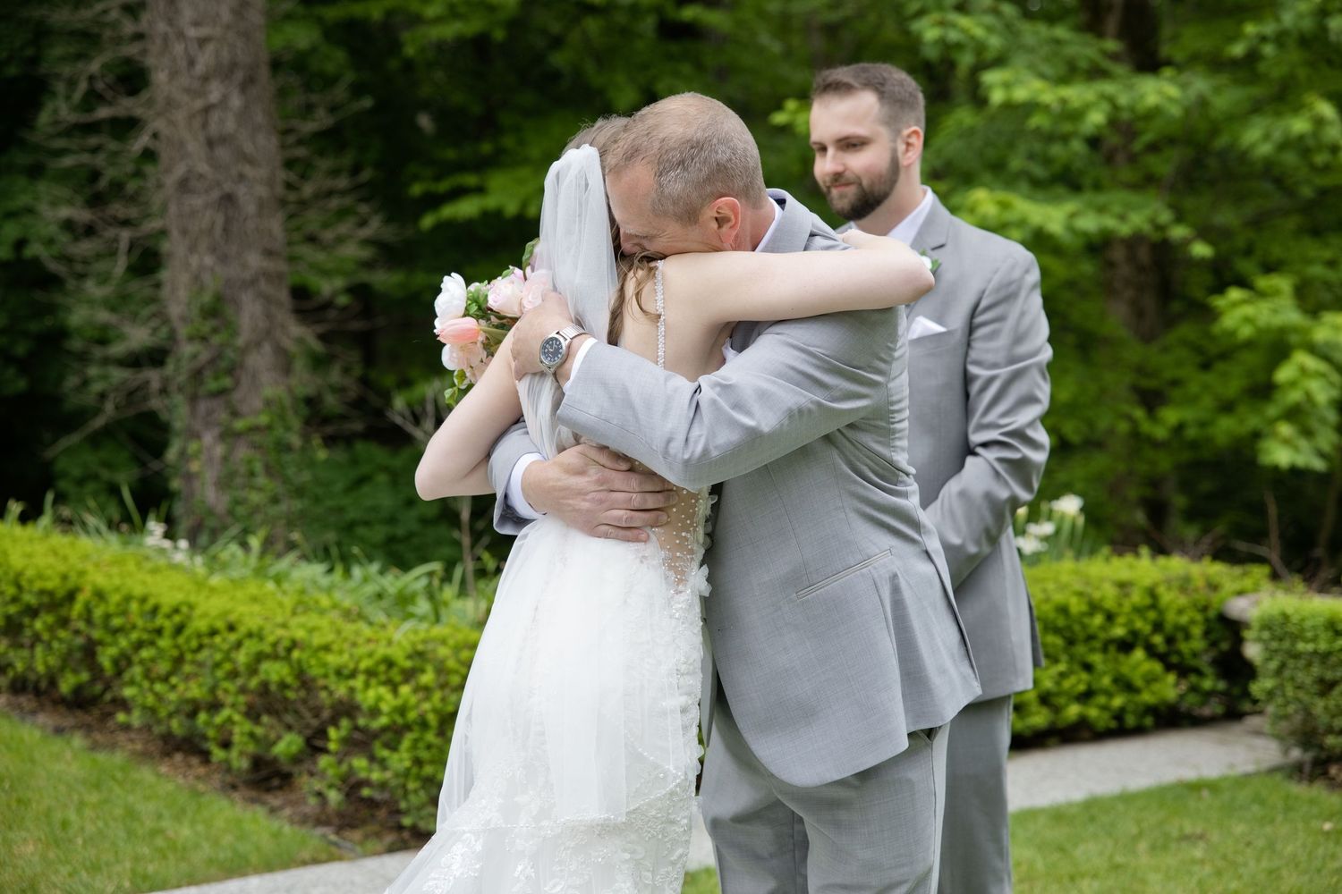 Bride in white lace gown hugging a guest while groom in gray suit smiles outdoors at a garden wedding.