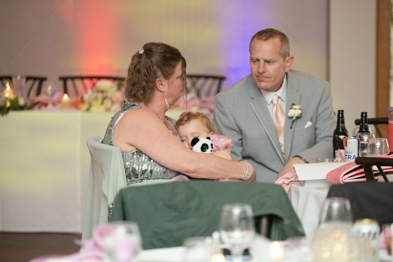 A woman holds a stuffed panda toy while conversing with a man in a suit at a wedding reception.