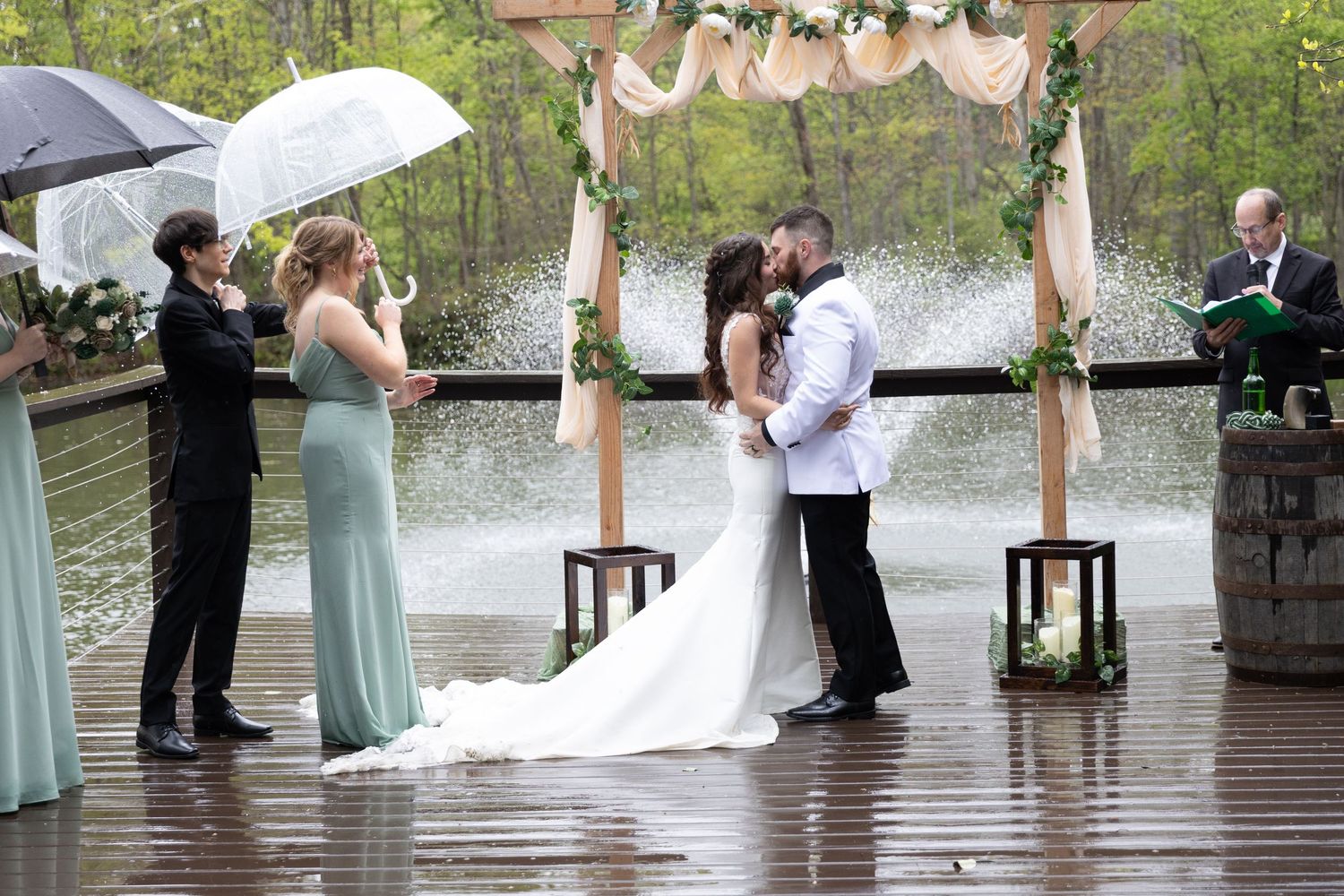 Bride and groom share first kiss at rainy outdoor wedding ceremony on wooden deck by a lake.