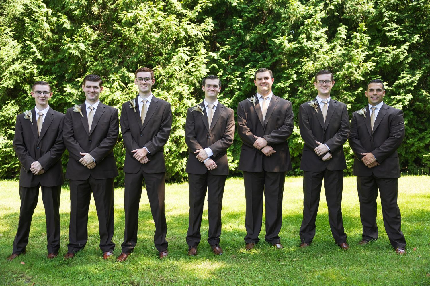Groomsmen in dark suits stand in line on green lawn.