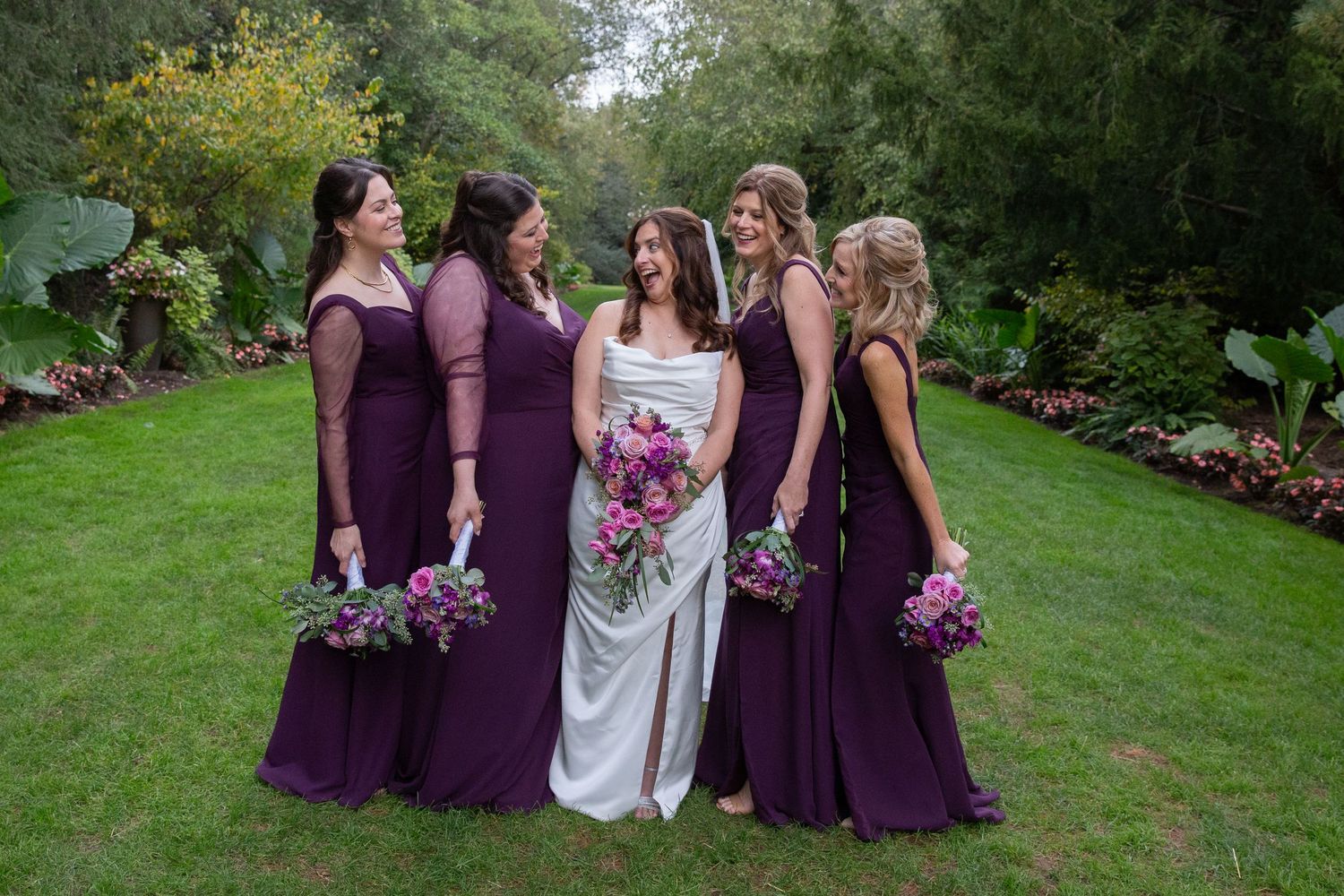 Wedding party members in purple dresses gathered outdoors in garden setting.