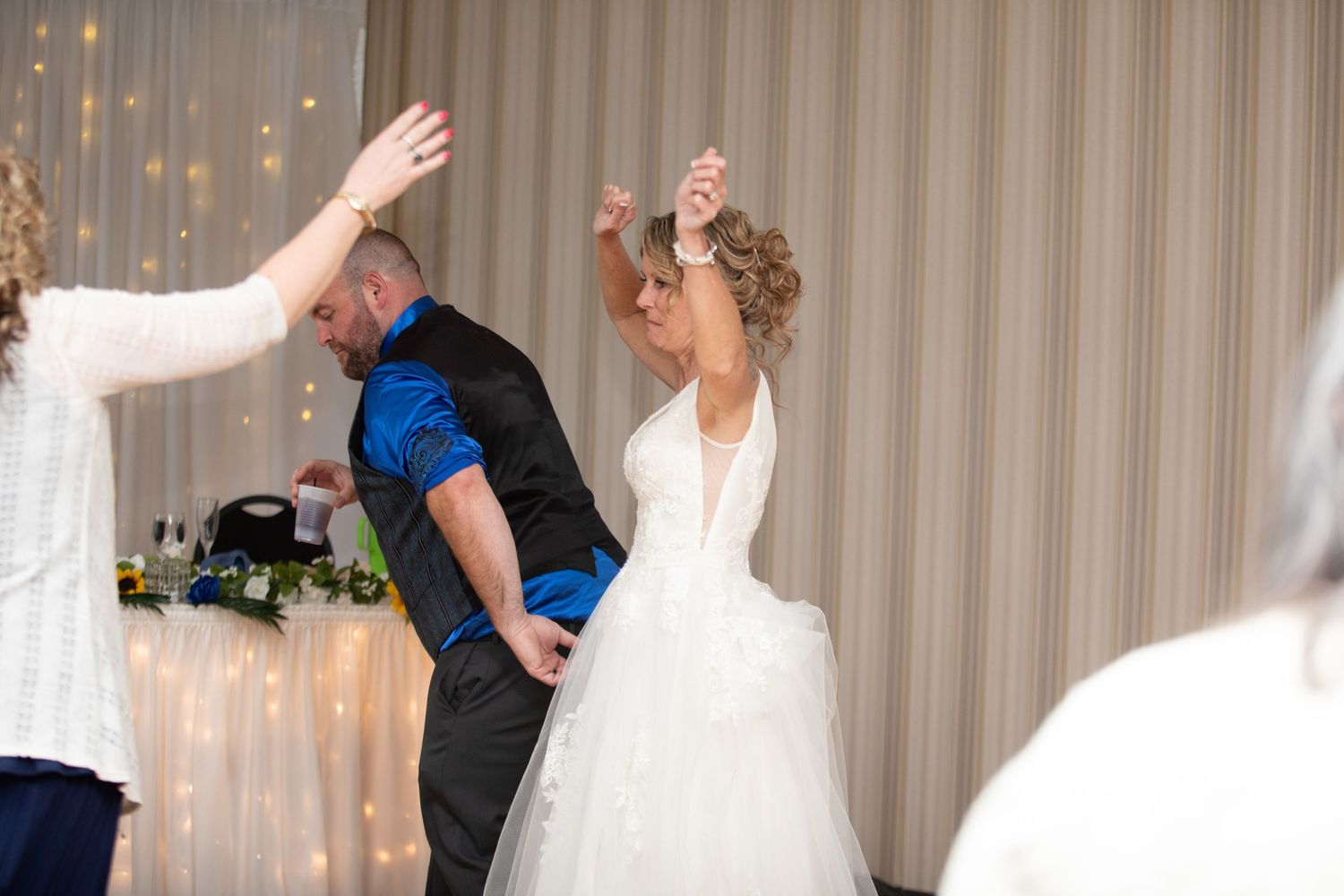 Bride in white gown dancing with groom in blue vest at elegant wedding reception with fairy lights.